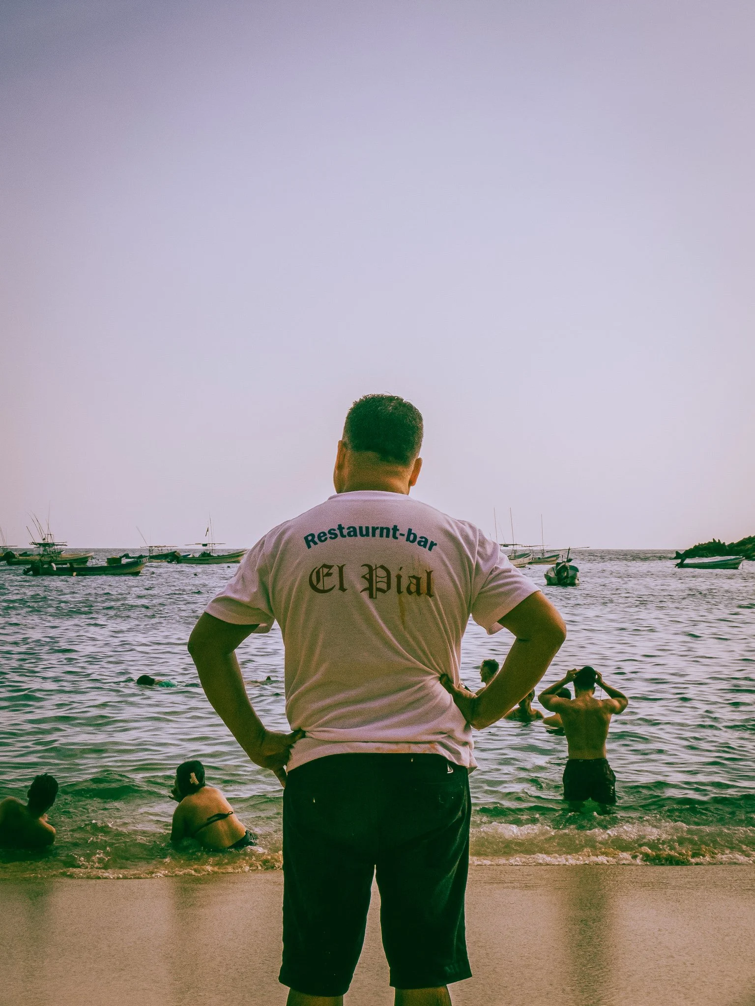 Man standing on beach with hands on hips, overlooking people swimming in the water, with boats in the distance, during sunset or sunrise.