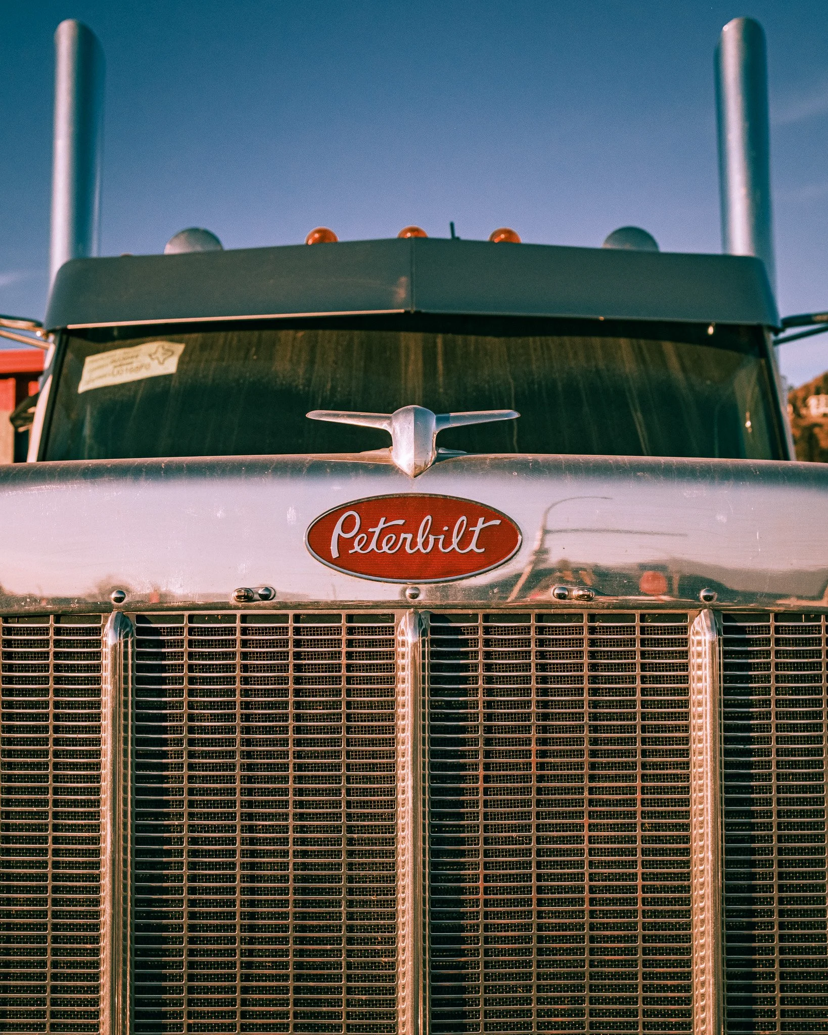 Front of Peterbilt semi-truck with a silver grille and a red oval Peterbilt logo, under a clear blue sky.