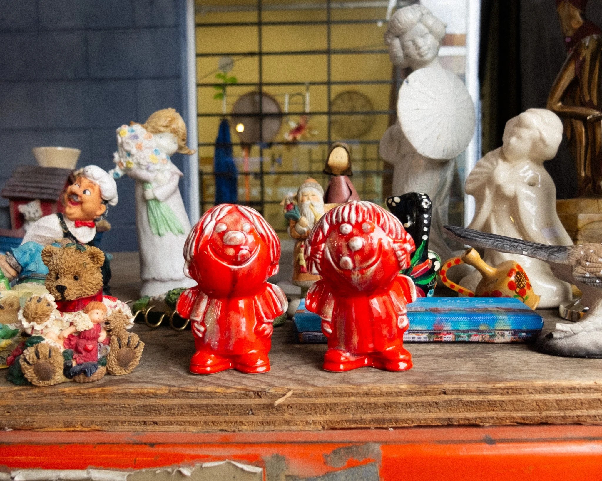 A collection of vintage figurines and knickknacks, including two red ceramic smiling gnome-like characters in the center, surrounded by various ceramic, plastic, and fabric figurines, some resembling children, animals, and mythical creatures, arranged on a wooden surface.