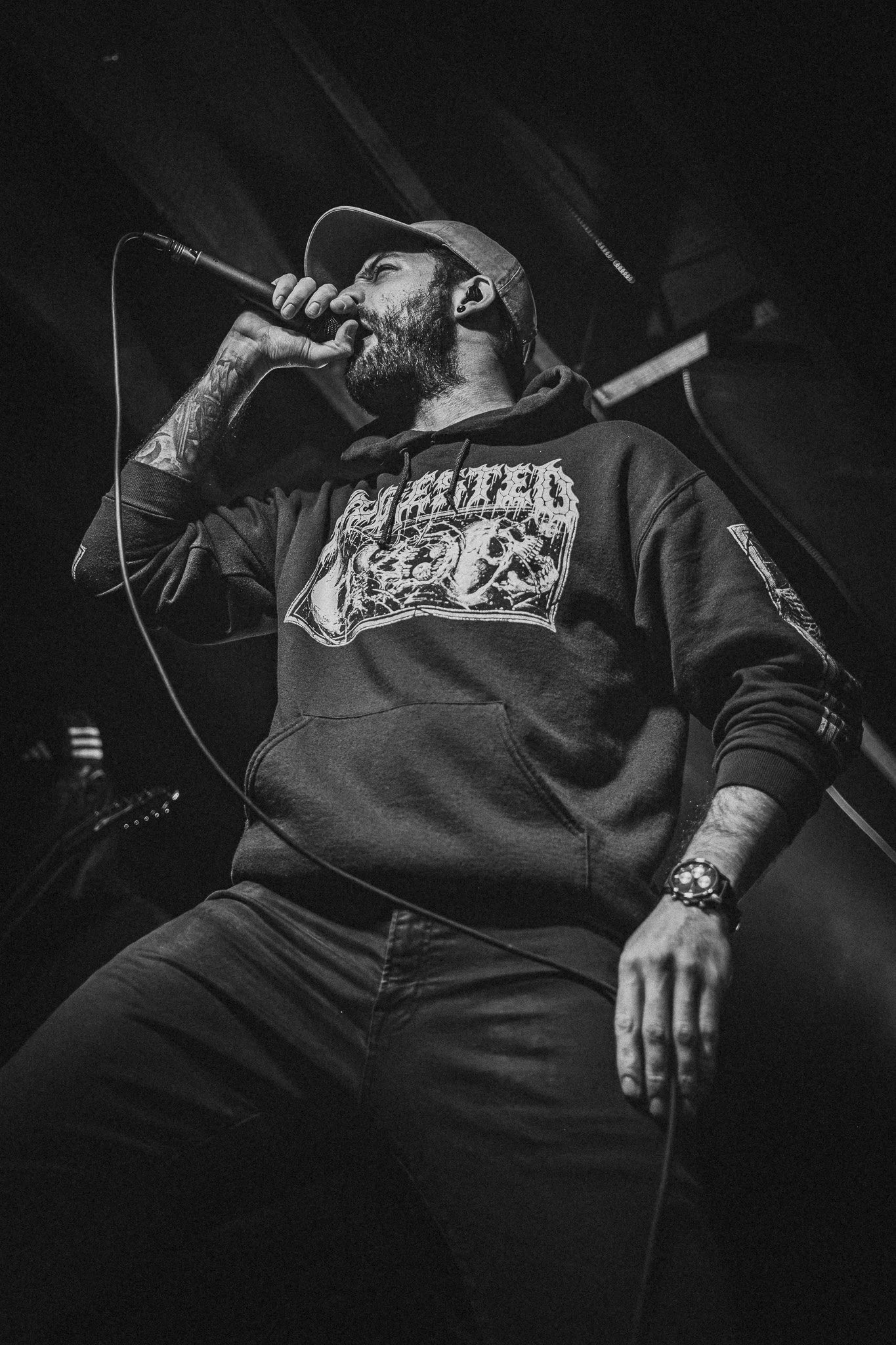 A man singing into a microphone on stage, wearing a hoodie, cap, and wristwatch in a dimly lit setting.