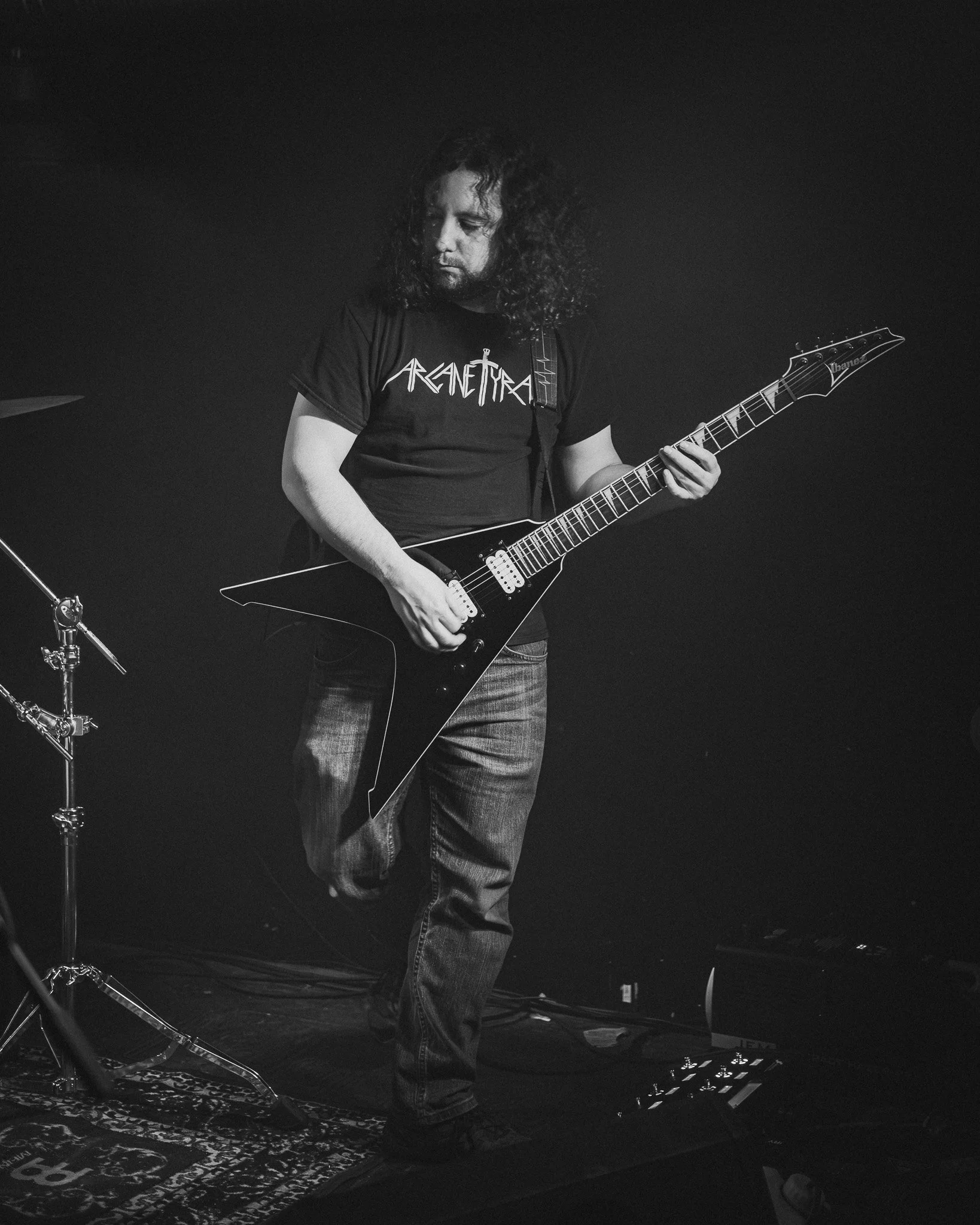 A male guitarist playing an electric guitar on stage, wearing a black 'Arch Enemy' t-shirt, jeans, and long hair, with a dark background and stage equipment.