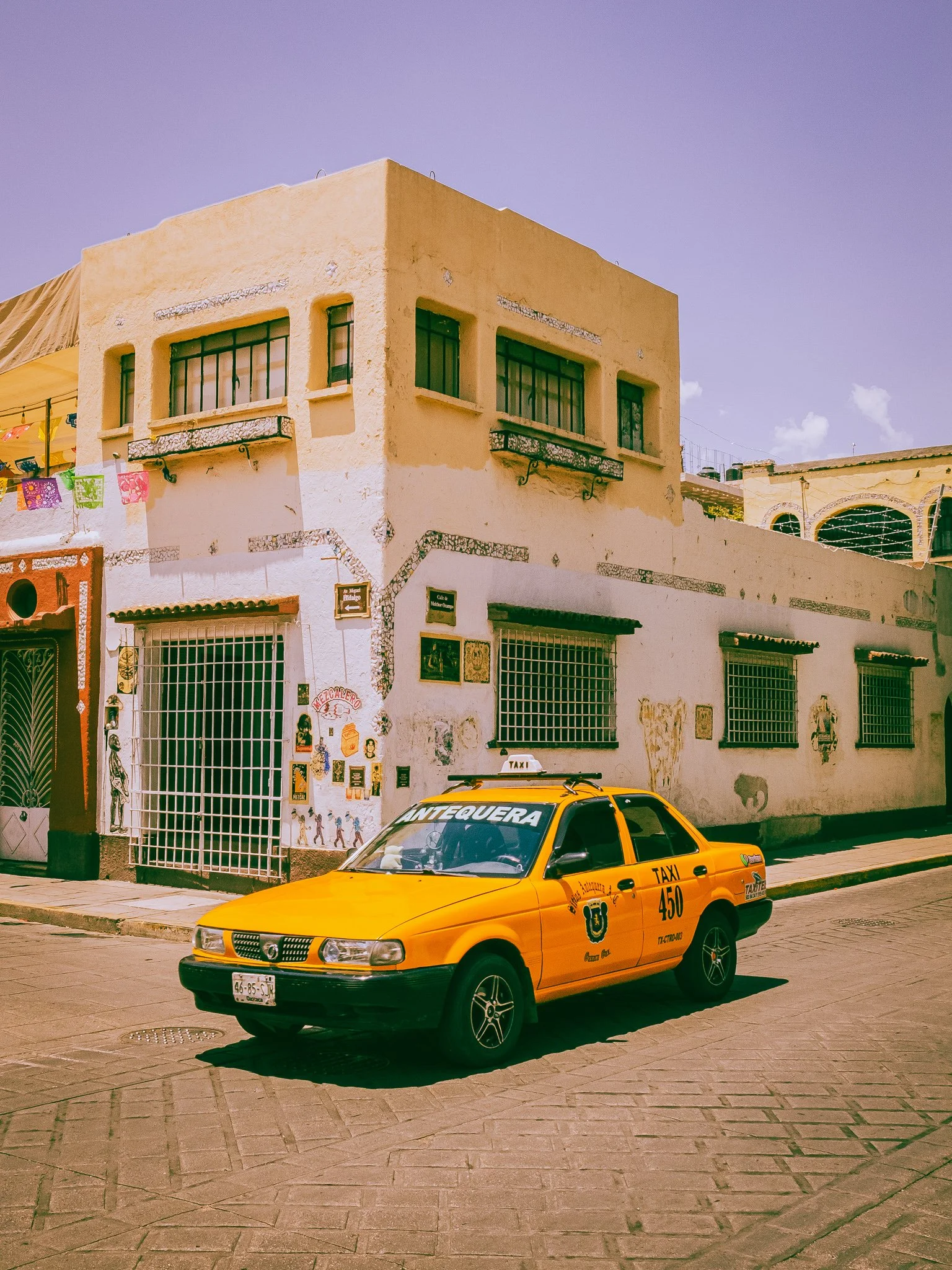 A bright yellow taxi parked on a street corner in front of a white building with barred windows, colorful decorations, and murals painted on the wall, under a clear sky.