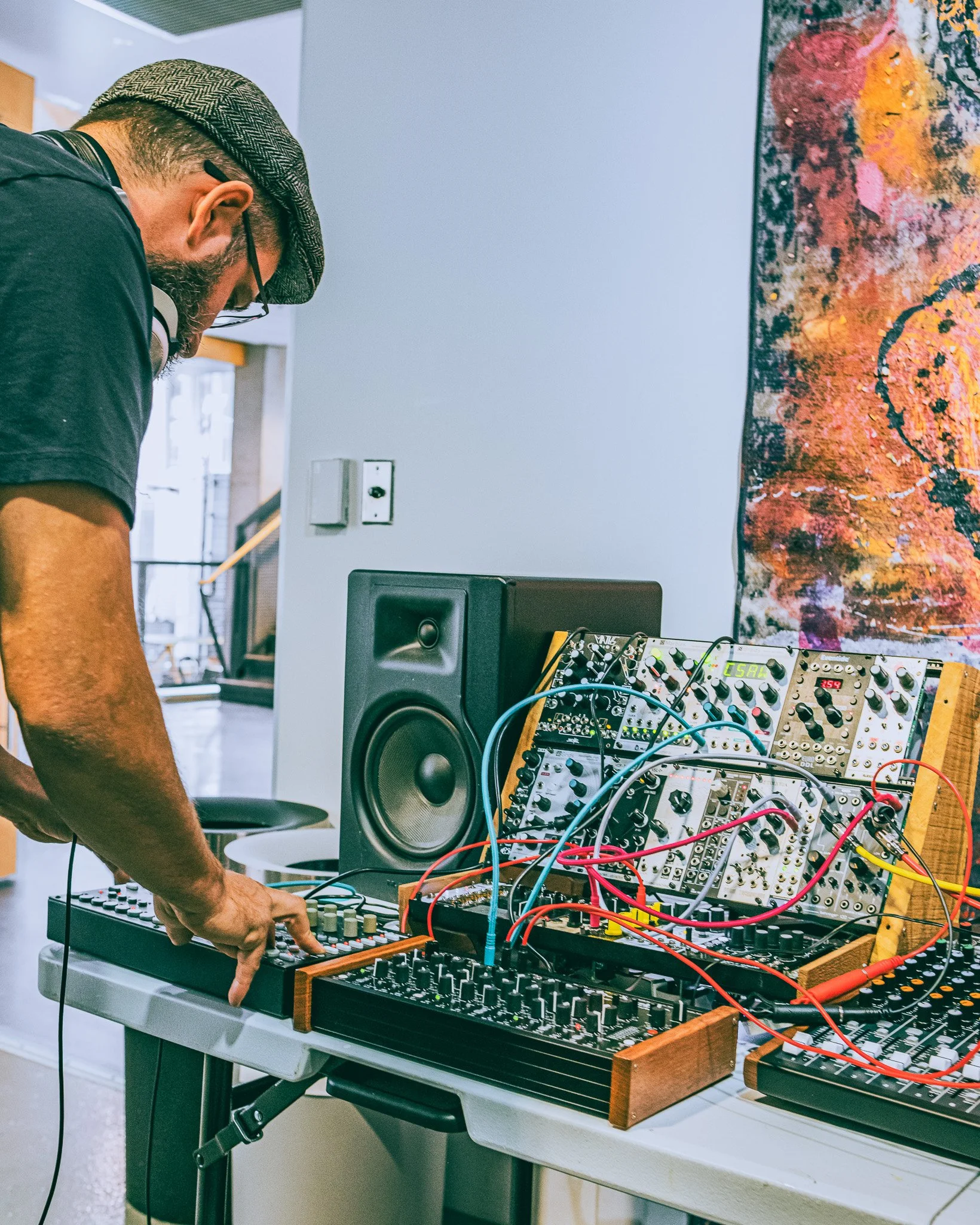 A person wearing a hat and glasses is operating a modular synthesizer and electronic music equipment on a table in an art studio.