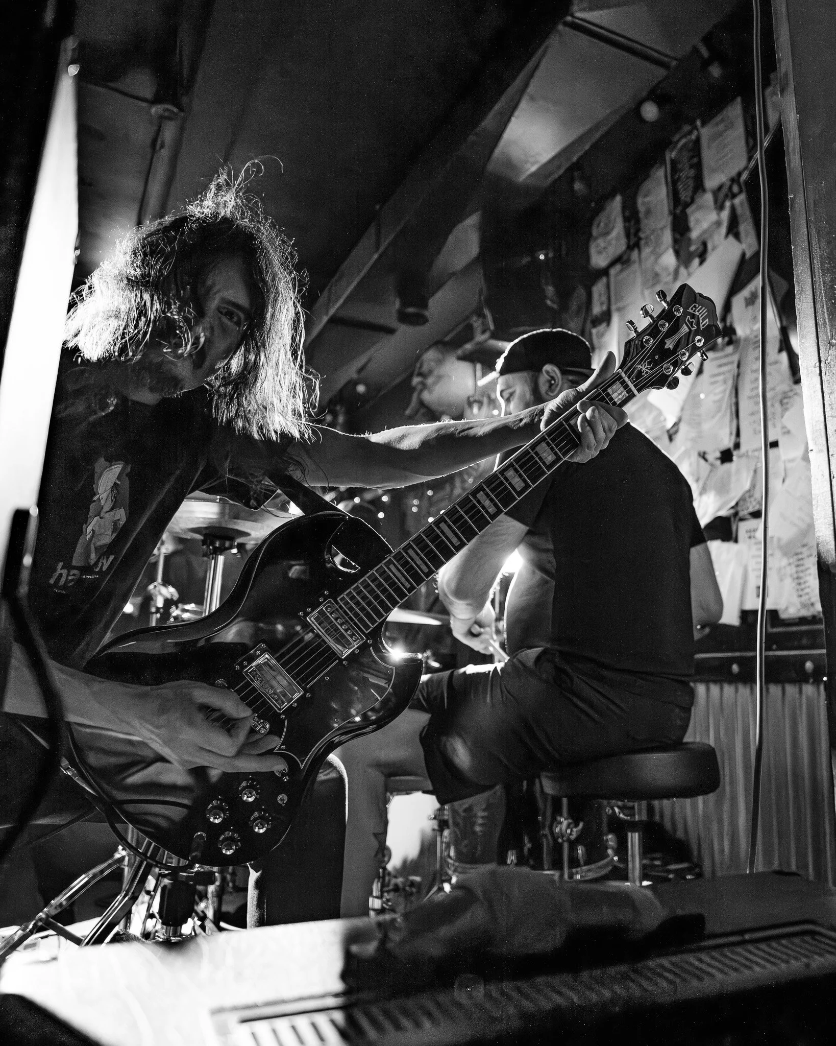 A musician with long, curly hair playing an electric guitar in a small, dimly lit venue with posters on the wall behind them.