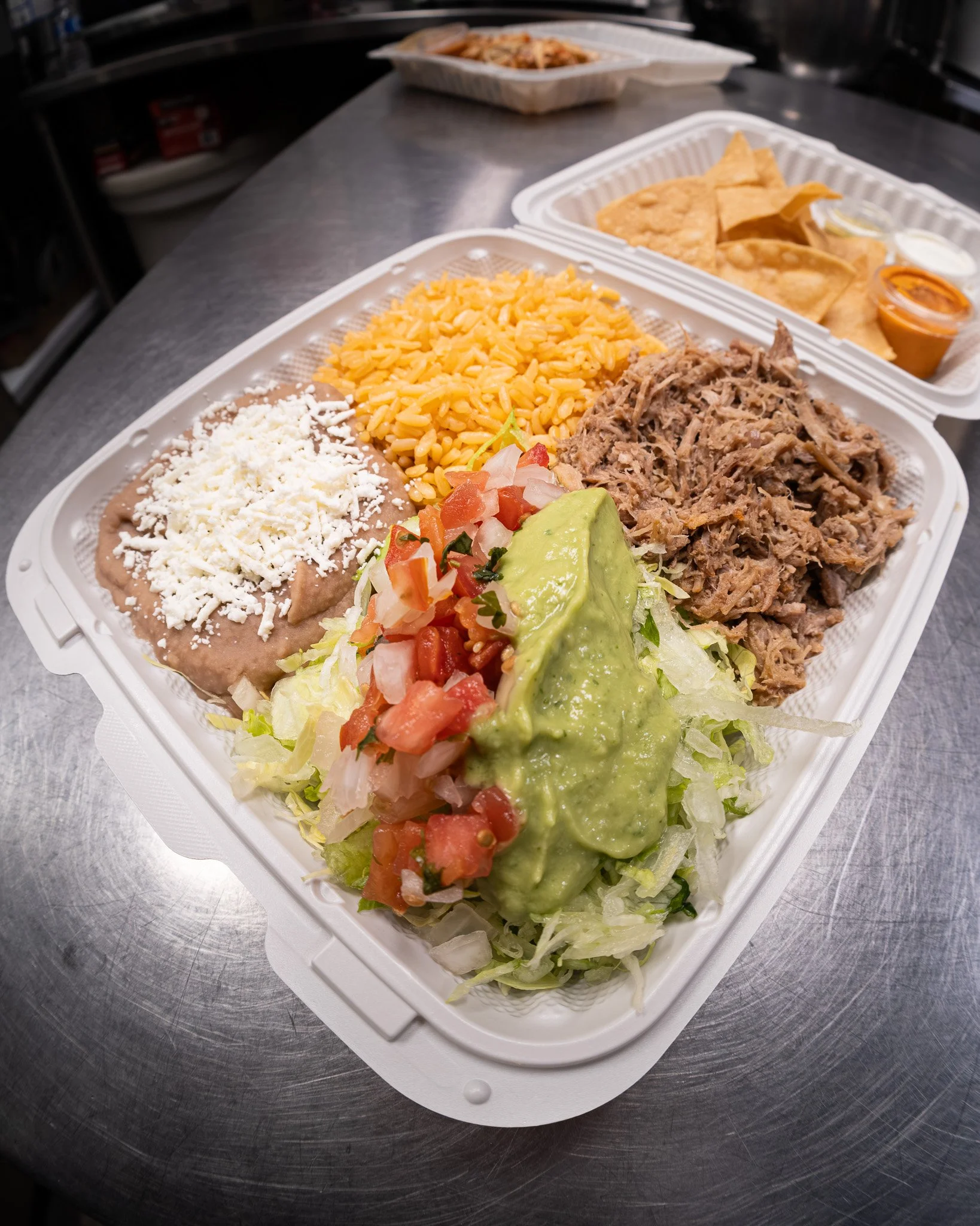 Mexican food platter with shredded beef, rice, refried beans topped with cheese, fresh salsa, lettuce, and guacamole, served with chips and sauces in the background.