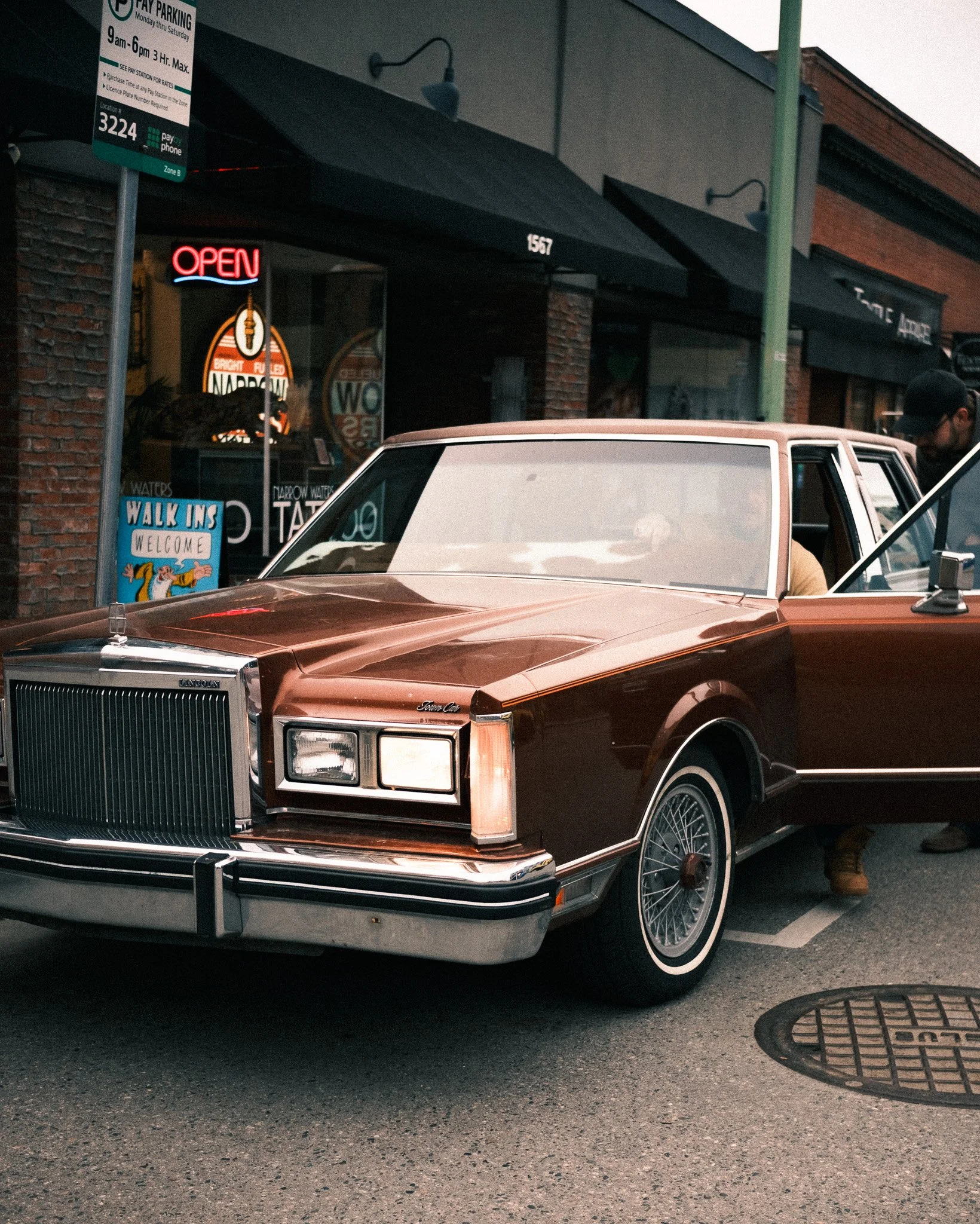 A vintage brown Lincoln Town Car parked on the street outside a storefront with an illuminated 'Open' sign. The car's driver is partially visible sitting inside. In the background, there is a sign indicating pay parking and a sidewalk sign welcoming 