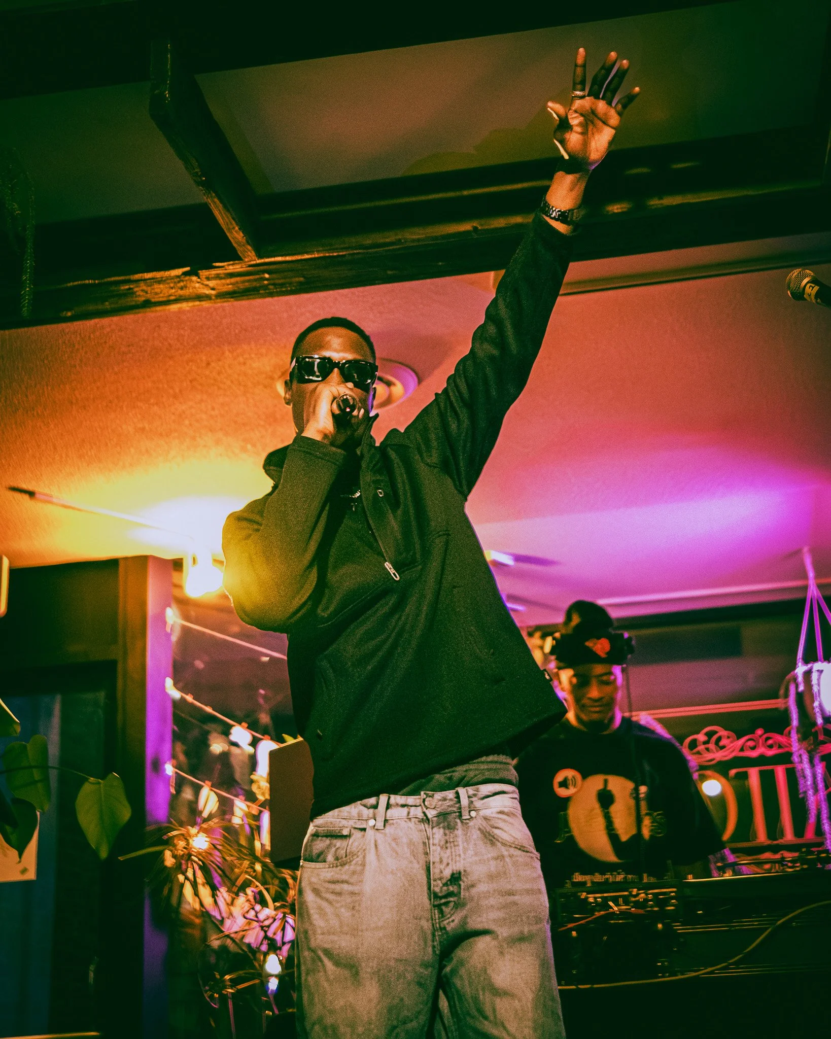 A man wearing sunglasses and a black jacket performs with a microphone on stage, raising his right hand, with a DJ in the background at a lively indoor event.