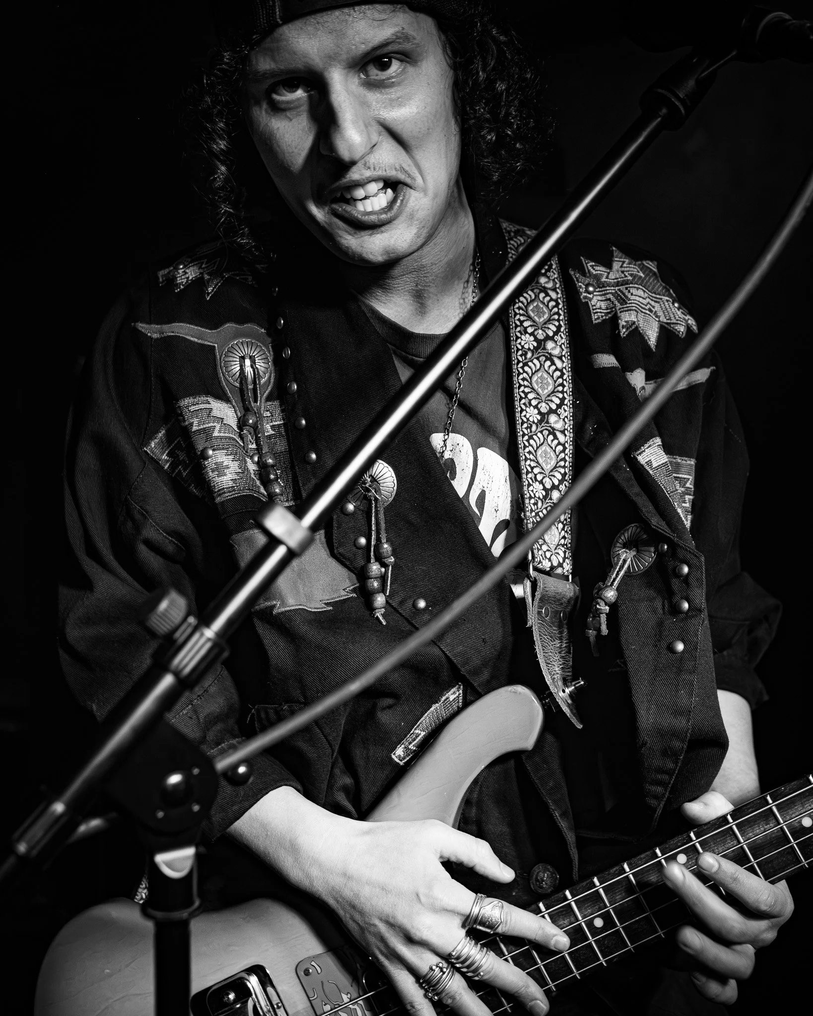 A person with curly hair and multiple rings, playing an electric guitar, singing into a microphone, wearing a decorated jacket, in a black and white photo.