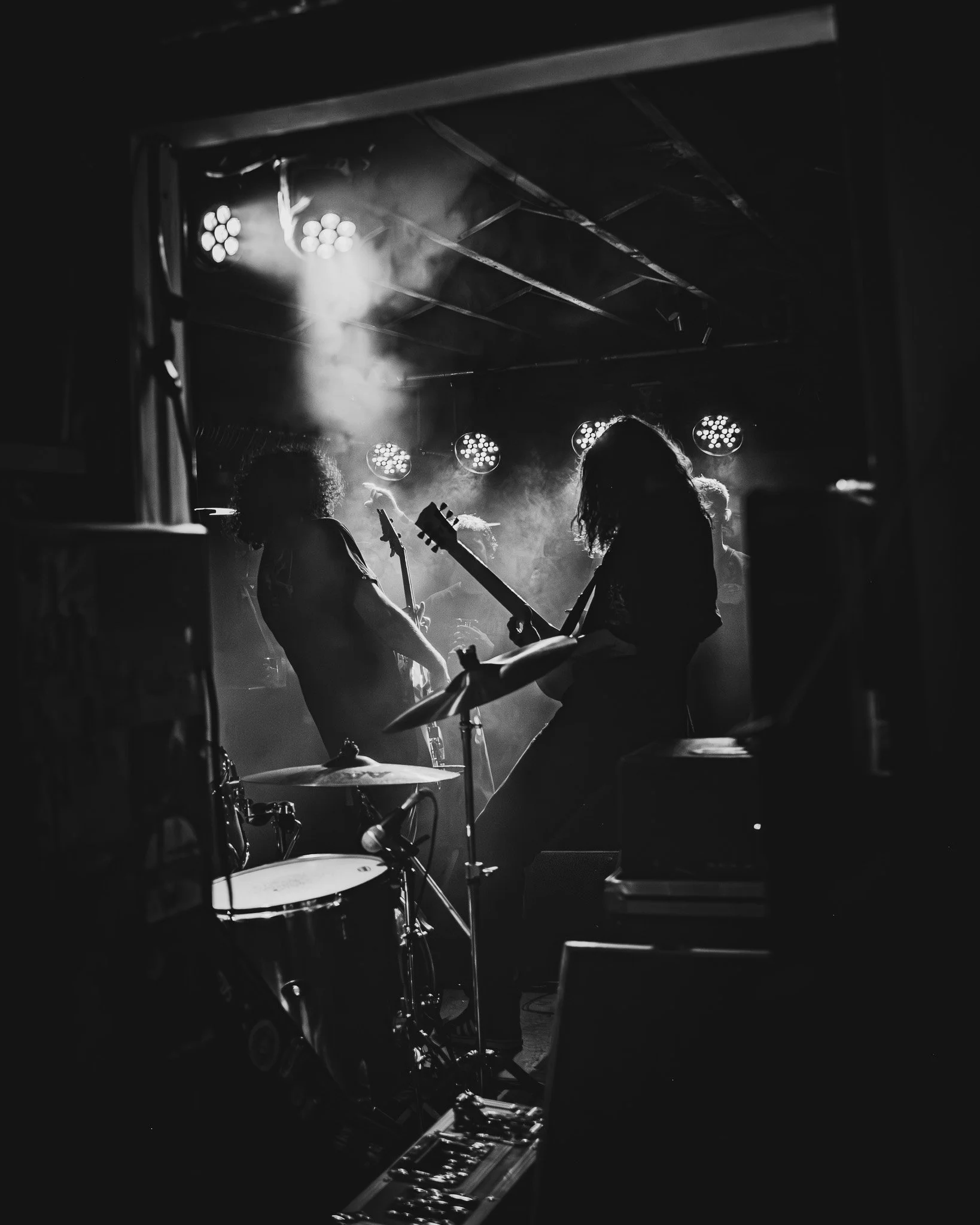 A black and white photo of a live band performing on stage, with silhouettes of musicians playing guitars and drums under stage lights, surrounded by fog.
