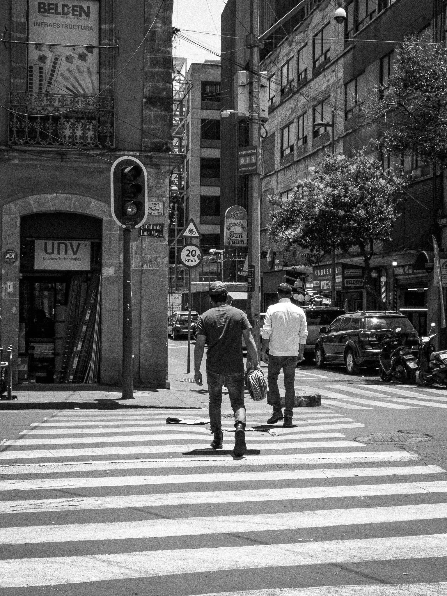 Two men walking across a zebra crossing in an urban area, with cars and motorcycles parked along the street and various buildings in the background.
