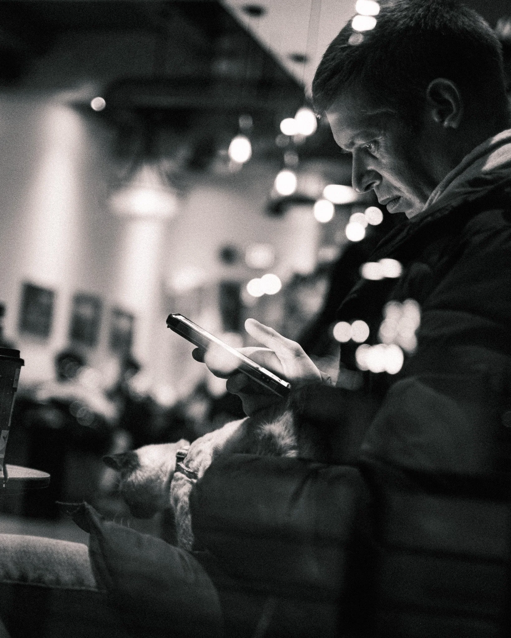 A man sitting in a public space looking at his phone, with a dog resting on his lap, in a black and white photo.