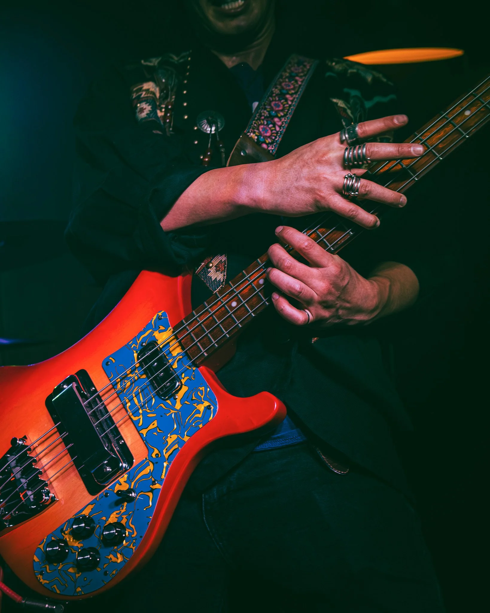 A person playing a colorful electric bass guitar, wearing multiple rings and a dark shirt with embroidered details.
