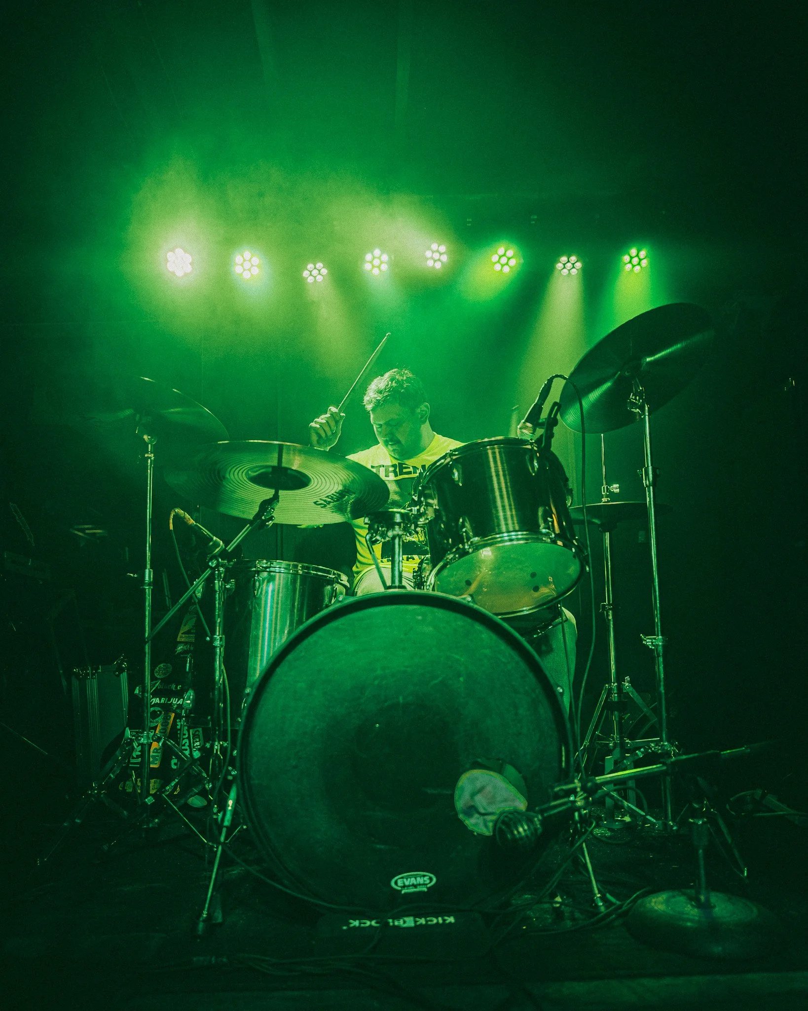 A drummer playing a drum kit on stage with green stage lighting.