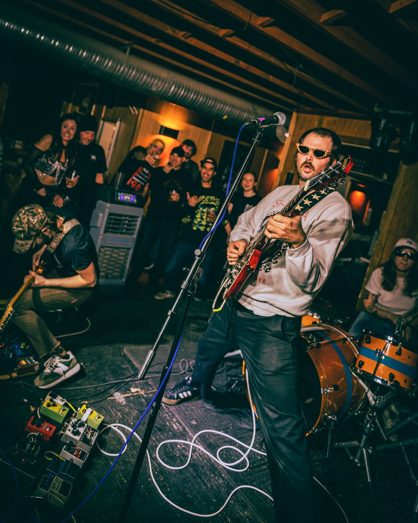 A band performs live music on stage with a guitarist wearing sunglasses, surrounded by a small audience in a cozy, dimly-lit venue.
