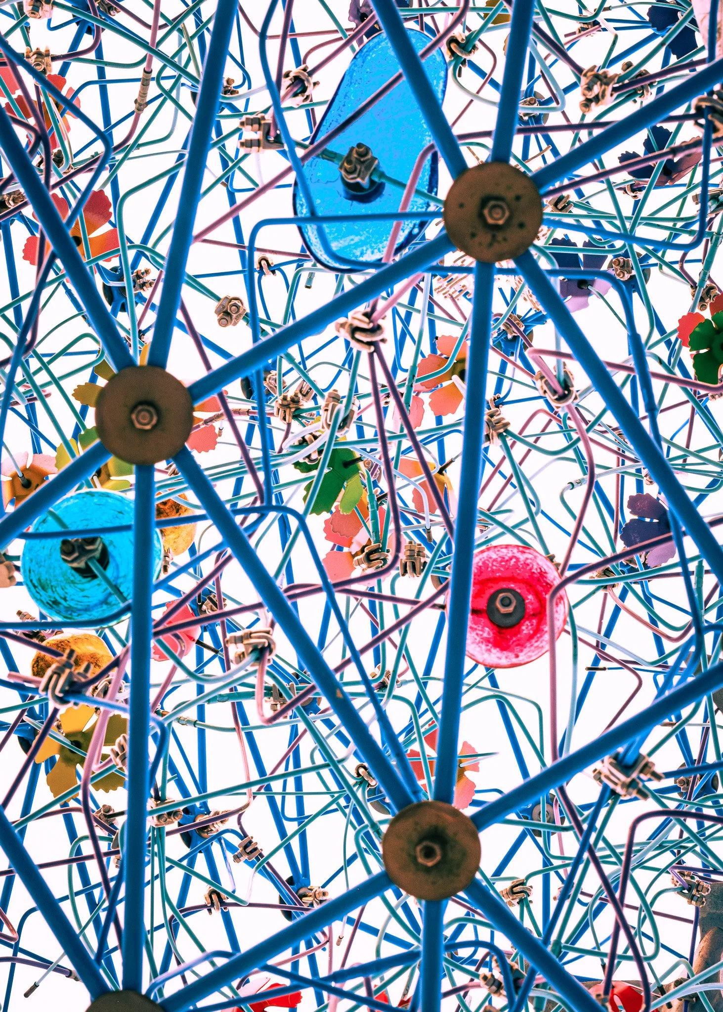 An intricate, colorful, abstract sculpture made of tangled wires, metal parts, and plastic elements, viewed from below against a white sky.