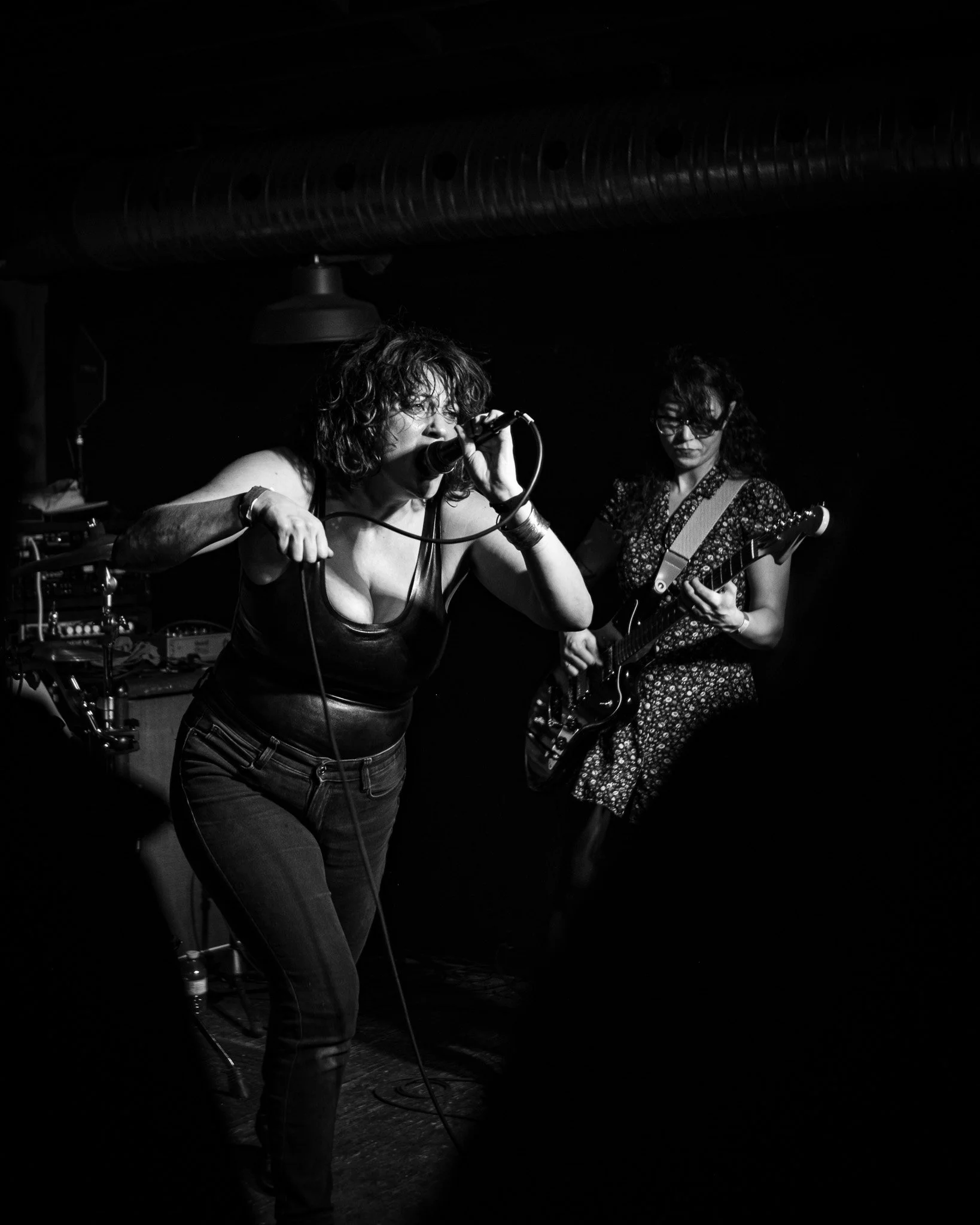 Black and white photo of two women performing on stage, one singing into a microphone and the other playing an electric guitar, in a dark setting.
