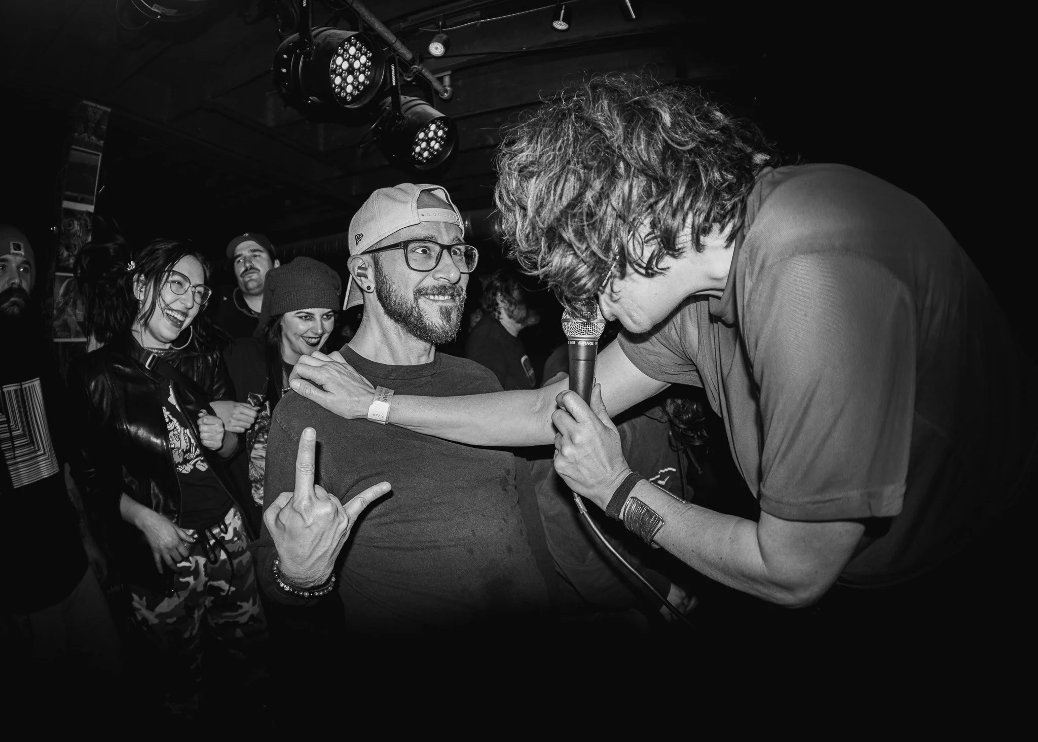 A man with curly hair leaning forward with a microphone toward a man with glasses, a beard, and a cap. The smiling man with glasses is making a rock hand gesture with his right hand. A group of people in the background watching and smiling.