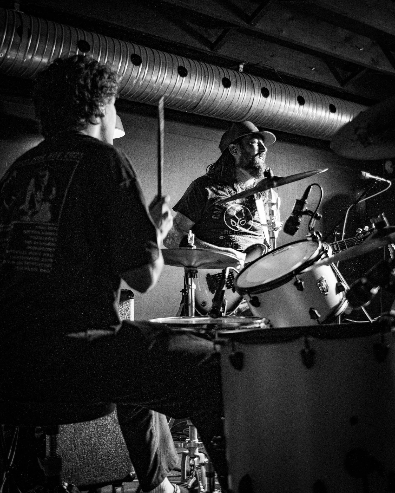 Two musicians playing drums on stage. The person on the left is sitting and holding drumsticks, and the person on the right has long hair, a beard, a cap, and is playing a snare drum. The scene is in black and white with a dark background.
