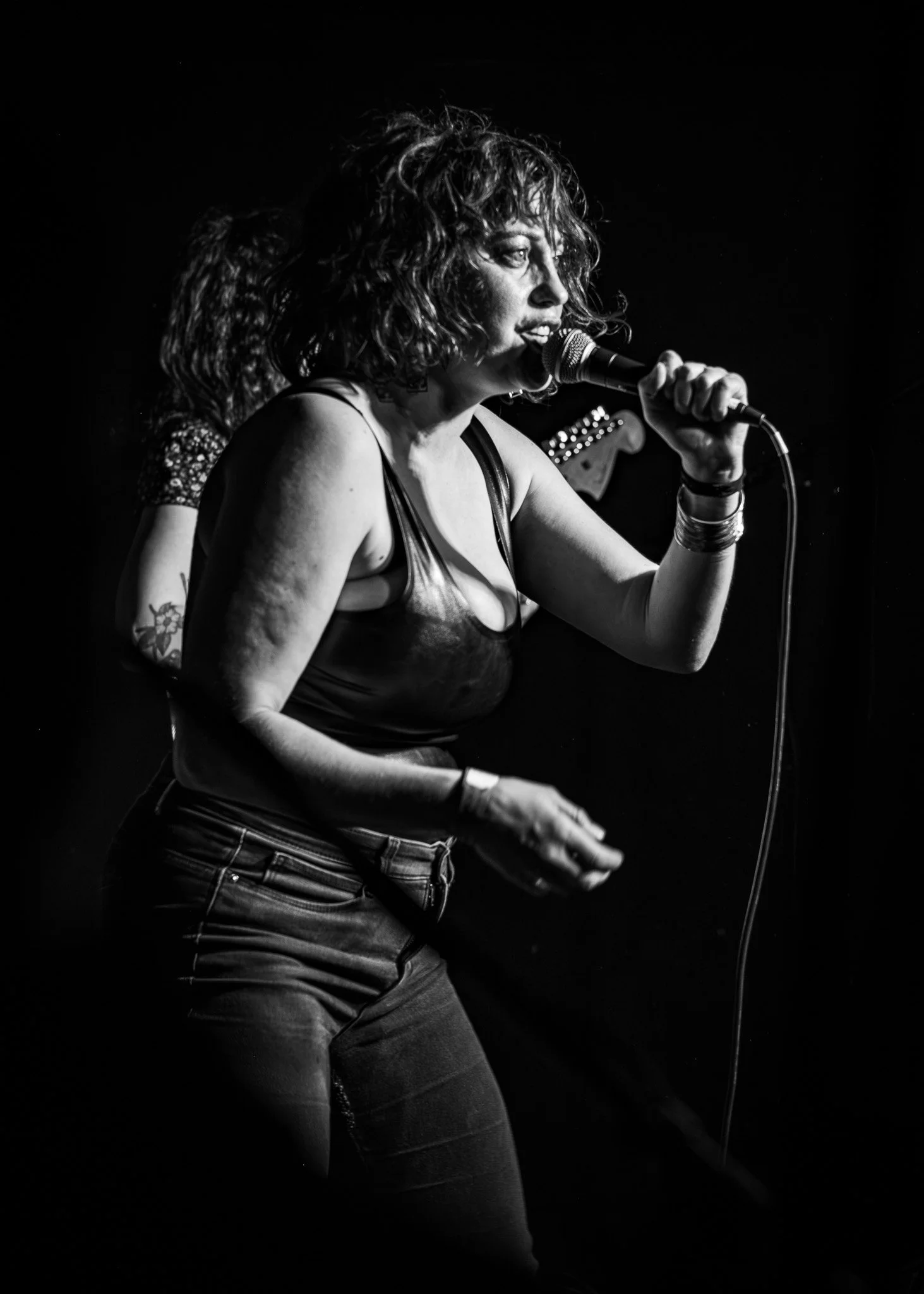 Black and white photo of a woman singing into a microphone on stage, with her curly hair and casual outfit visible.
