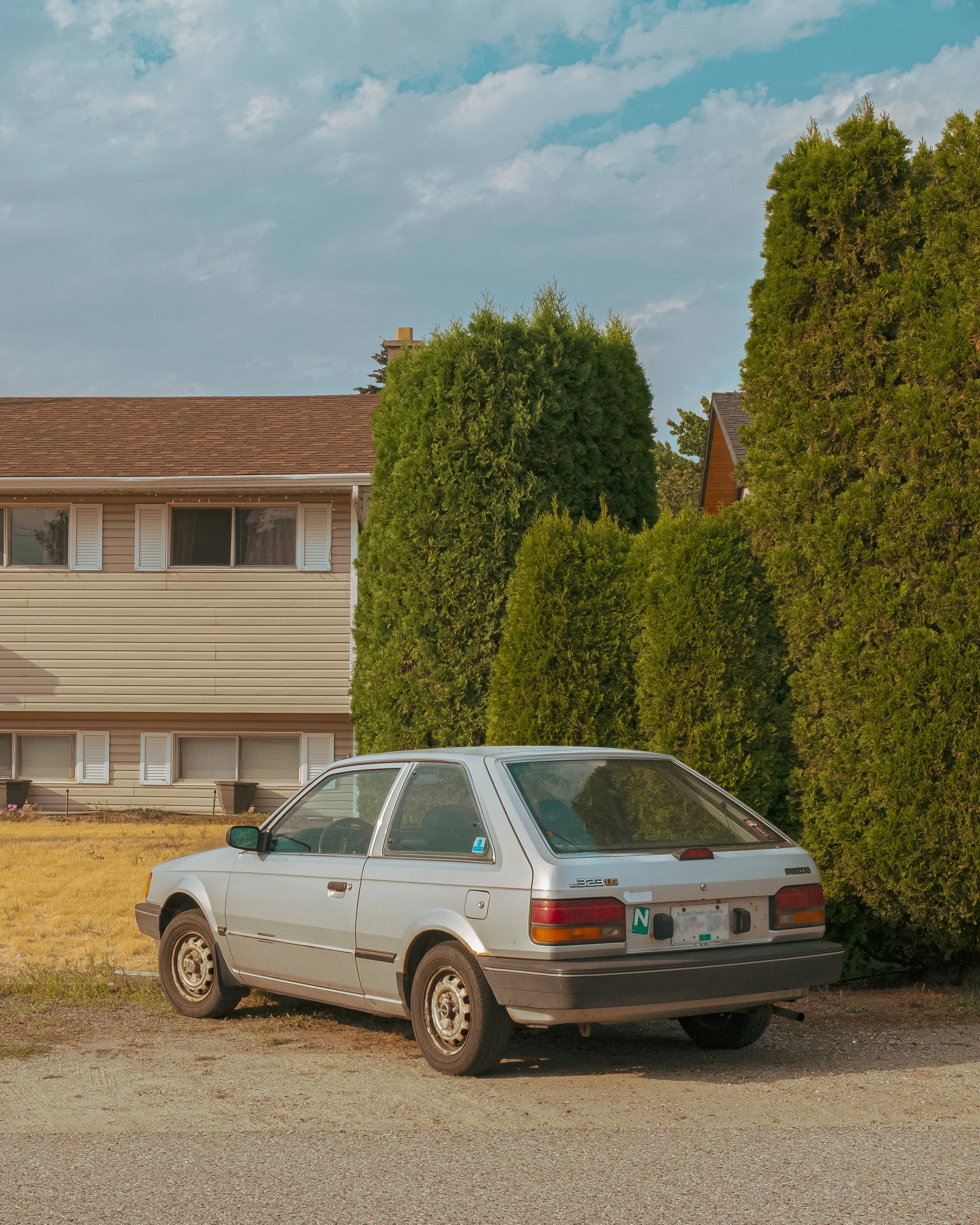 A silver, vintage hatchback car parked on a dirt driveway in front of a beige house with white shutters and overgrown greenery.