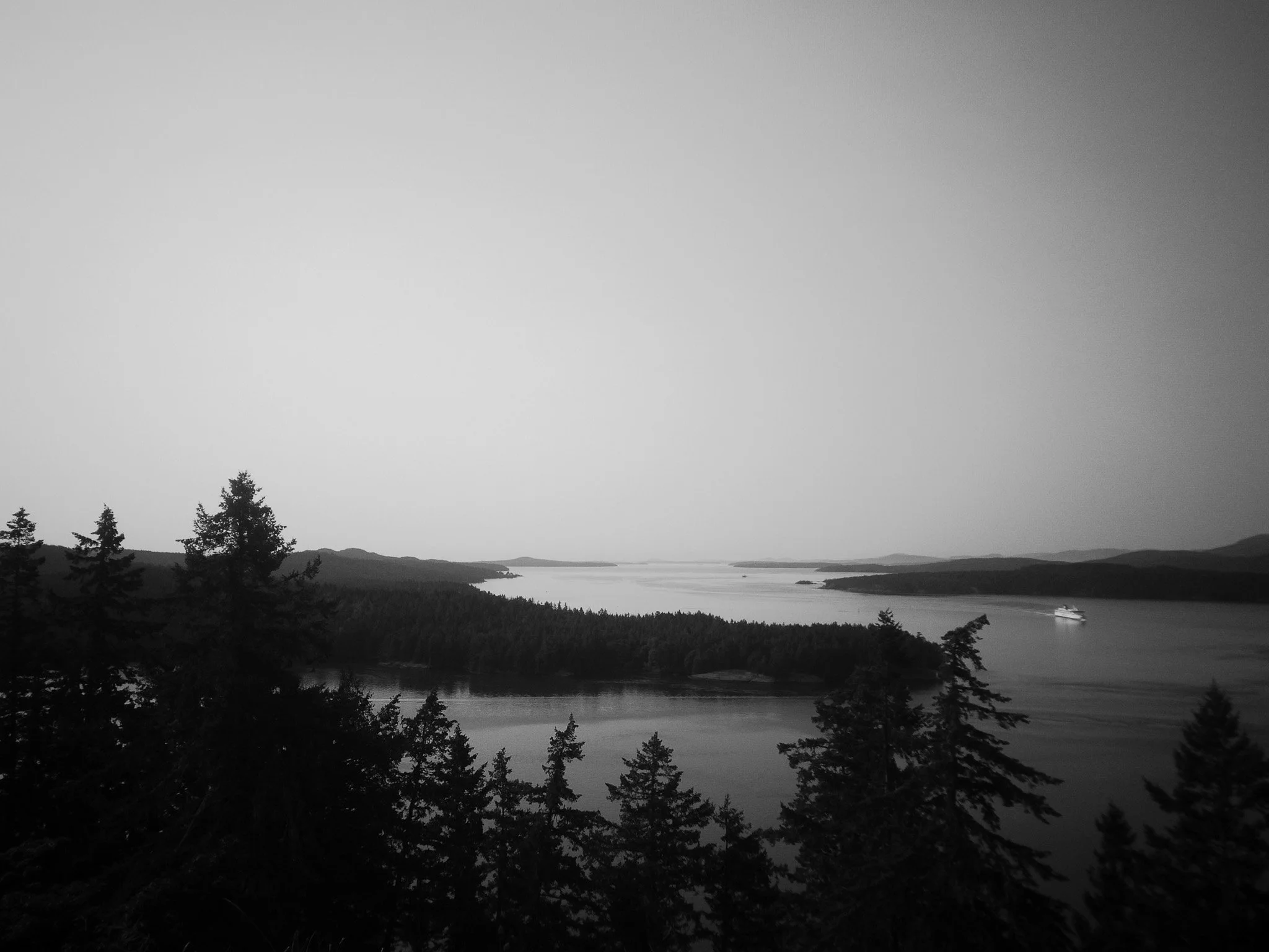 A black and white landscape of a large body of water with small islands and a boat in the distance, surrounded by forested hills and mountains.