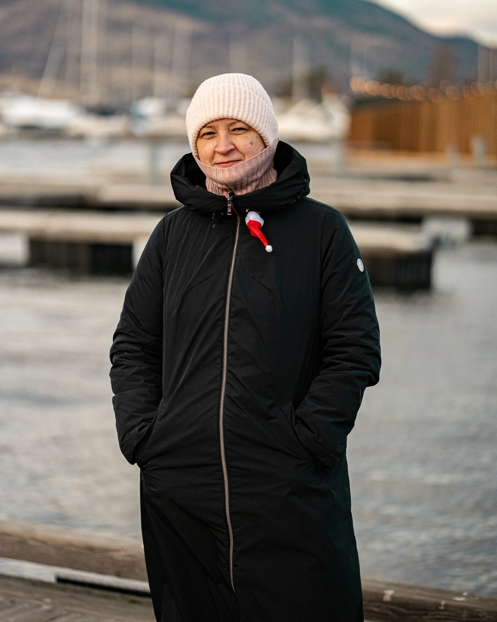 A woman dressed in a black winter coat, pink knit hat, and pink scarf standing outdoors near a dock with boats in the background.