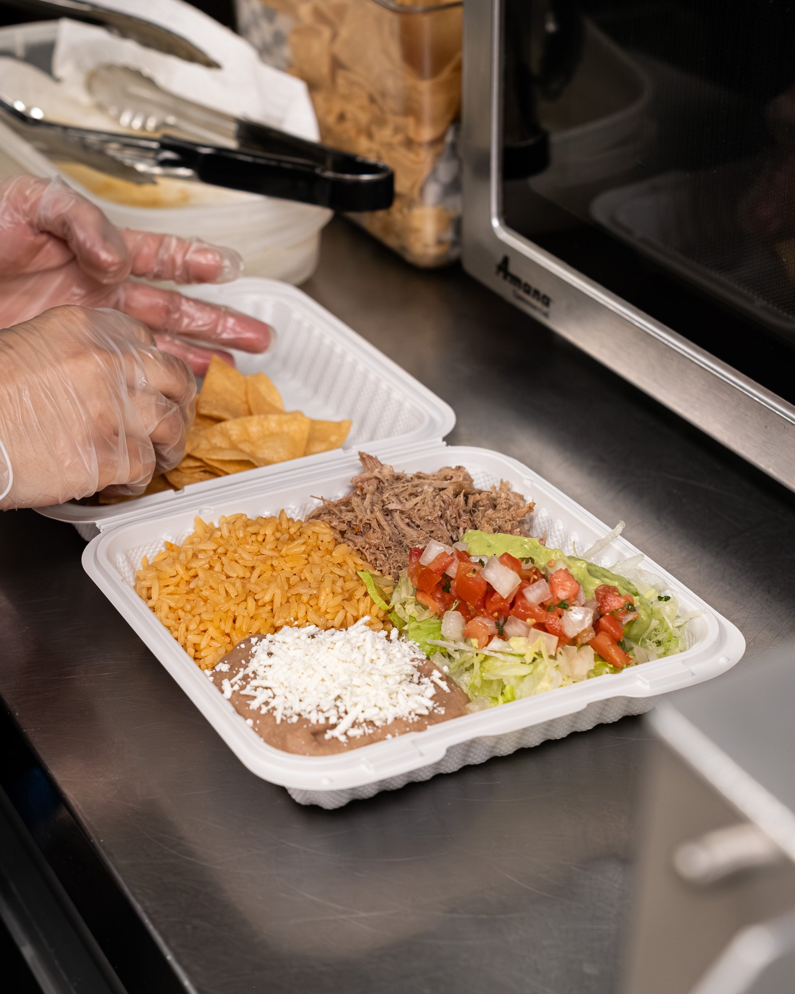 A takeout container with Mexican food including rice, shredded beef, refried beans topped with cheese, fresh lettuce and tomato salad, and tortilla chips.