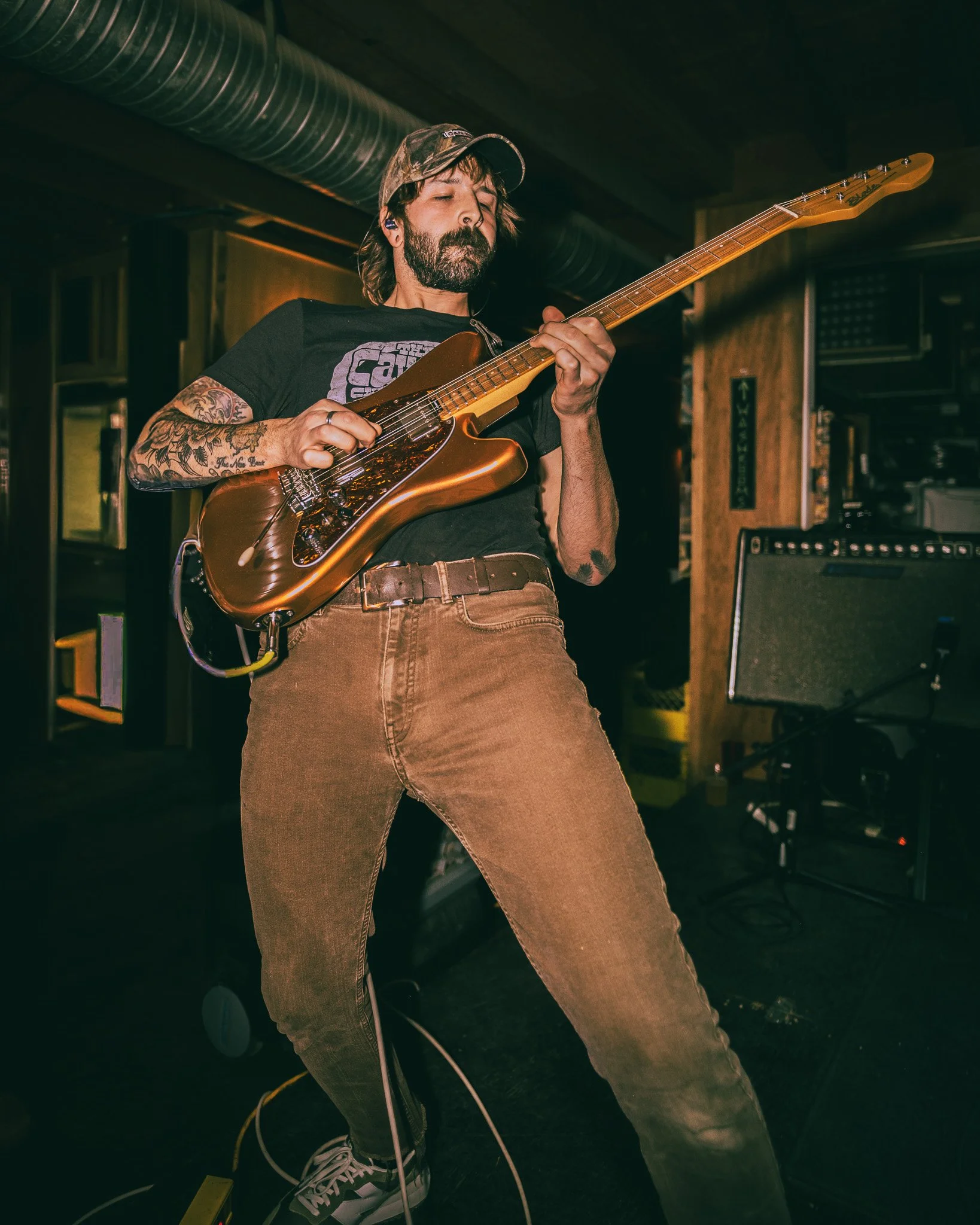 A man with tattoos, wearing a camouflage hat, black T-shirt, and brown pants, playing an electric guitar in a dimly lit indoor setting with wood-paneled walls.