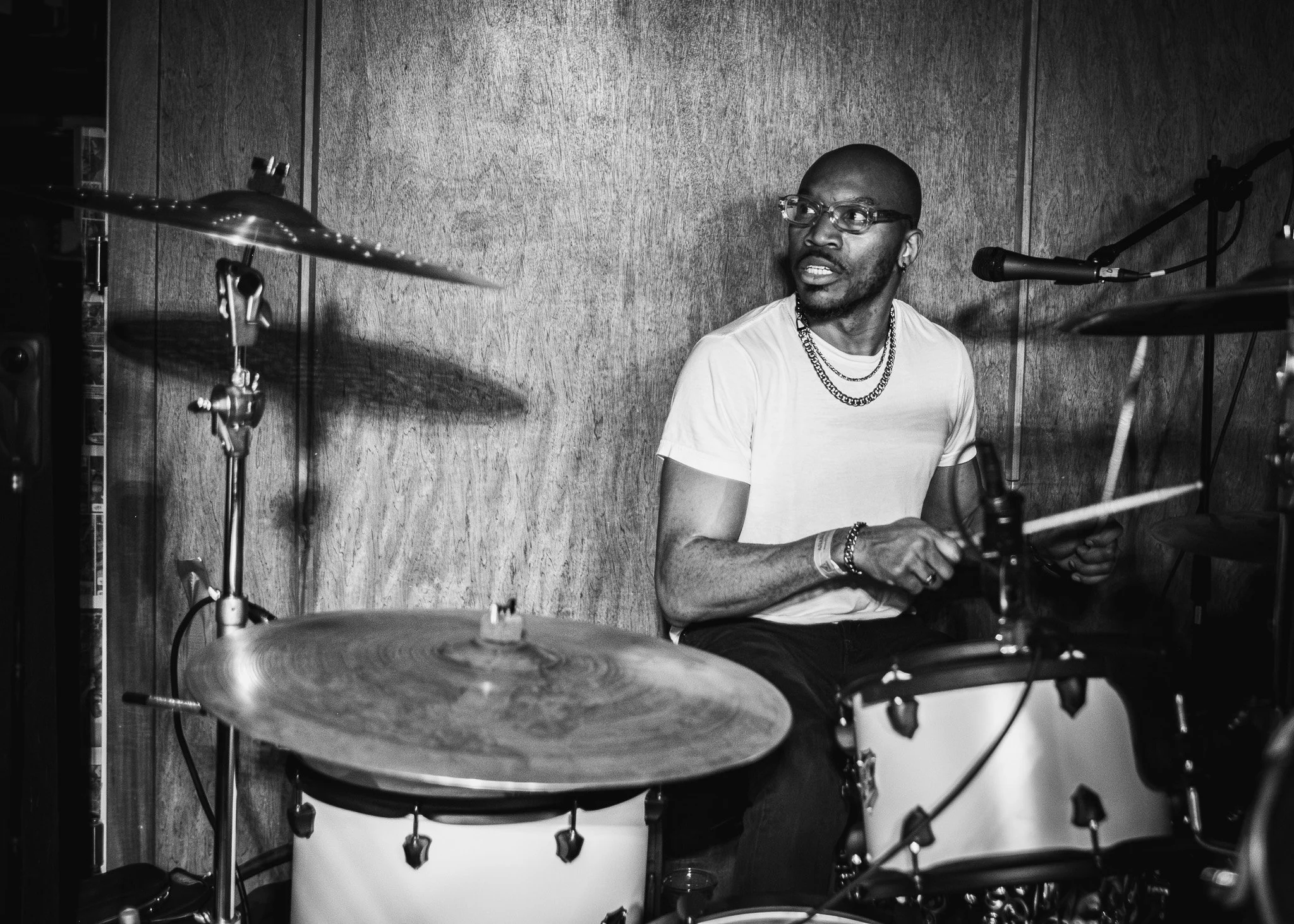 A Black man playing the drums in a music studio, wearing glasses and a white t-shirt, with a wood-paneled wall in the background.