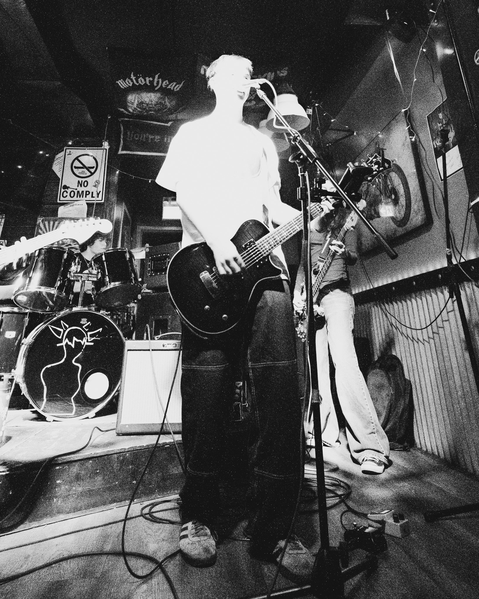 A black and white photo of a band performing on stage at a bar or club. The lead singer is playing an acoustic guitar and singing into a microphone, with a drum set and another guitarist in the background. There is a "No Smoking" sign on the wall and