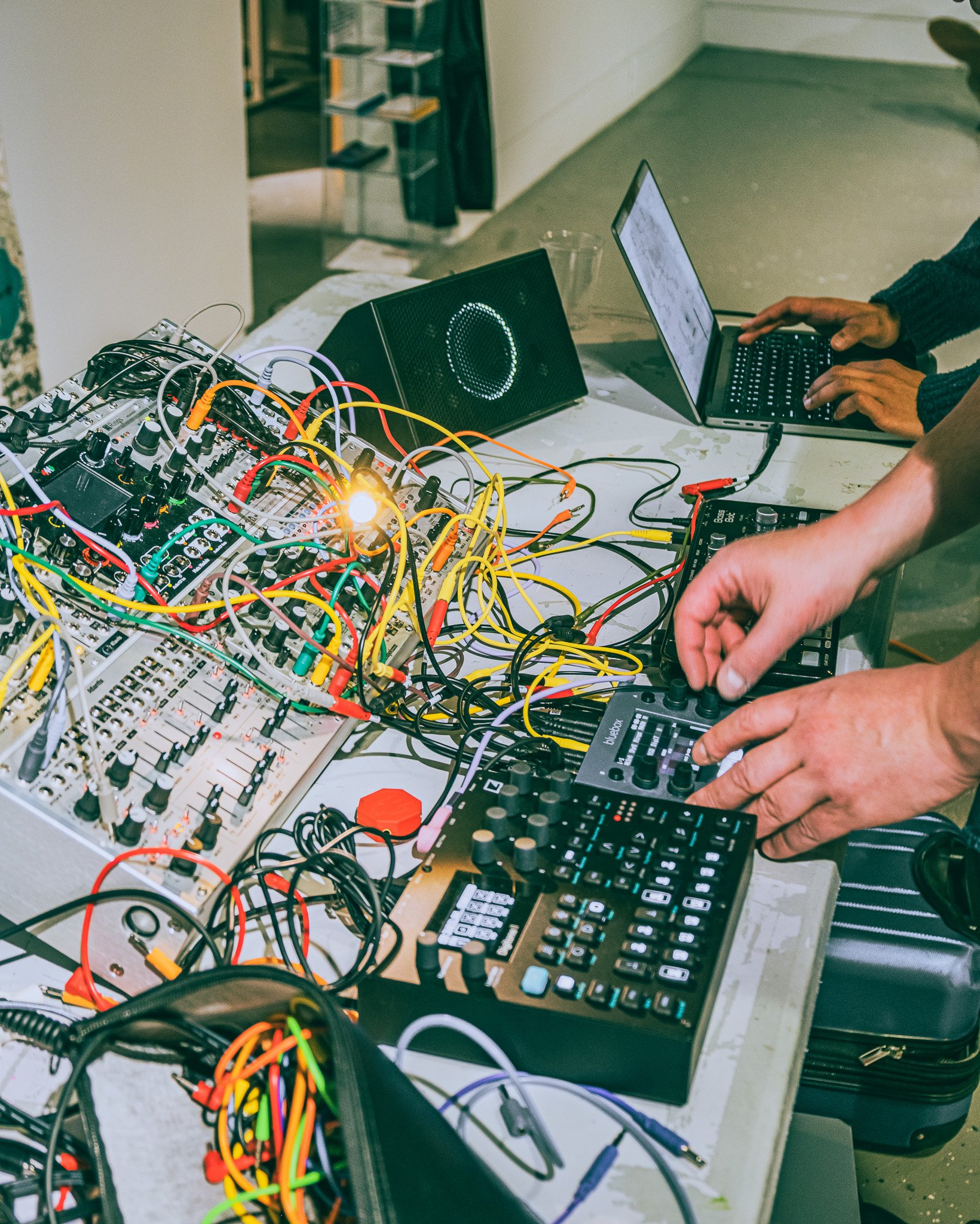 People working with electronic music equipment, including a synthesizer, mixer, and laptop, with colorful cables connecting the devices.