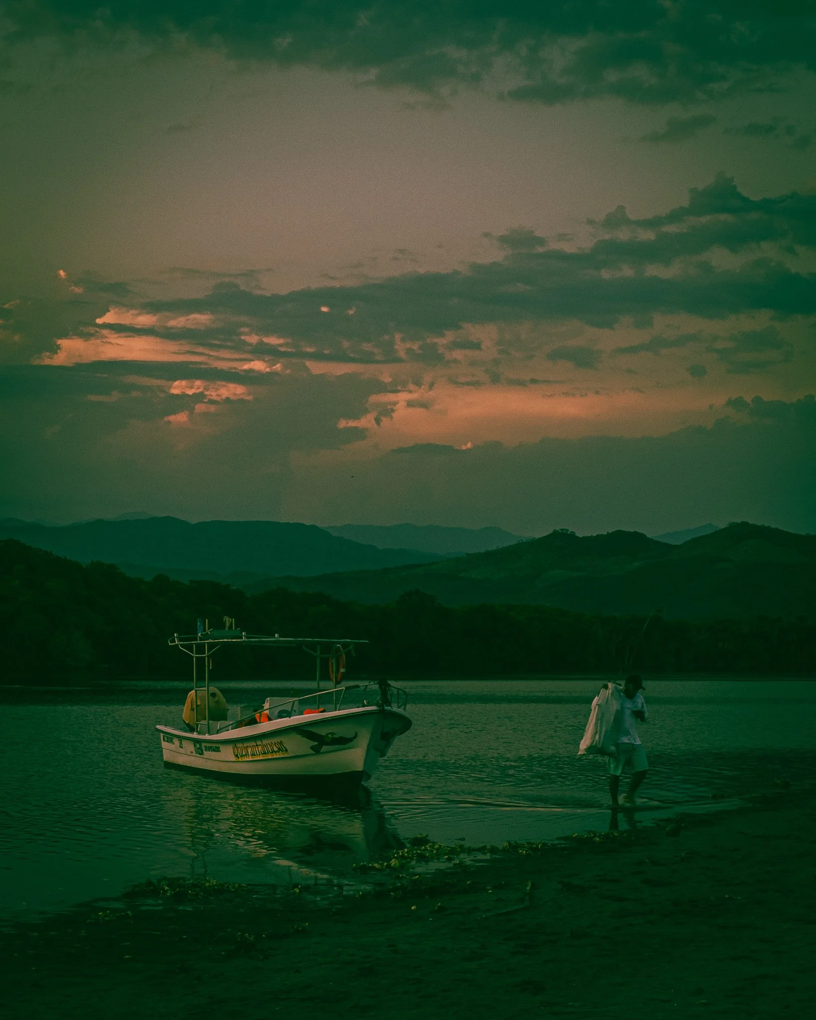 A boat anchored near the shore of a lake with mountains in the background and a person walking on the beach at sunset.