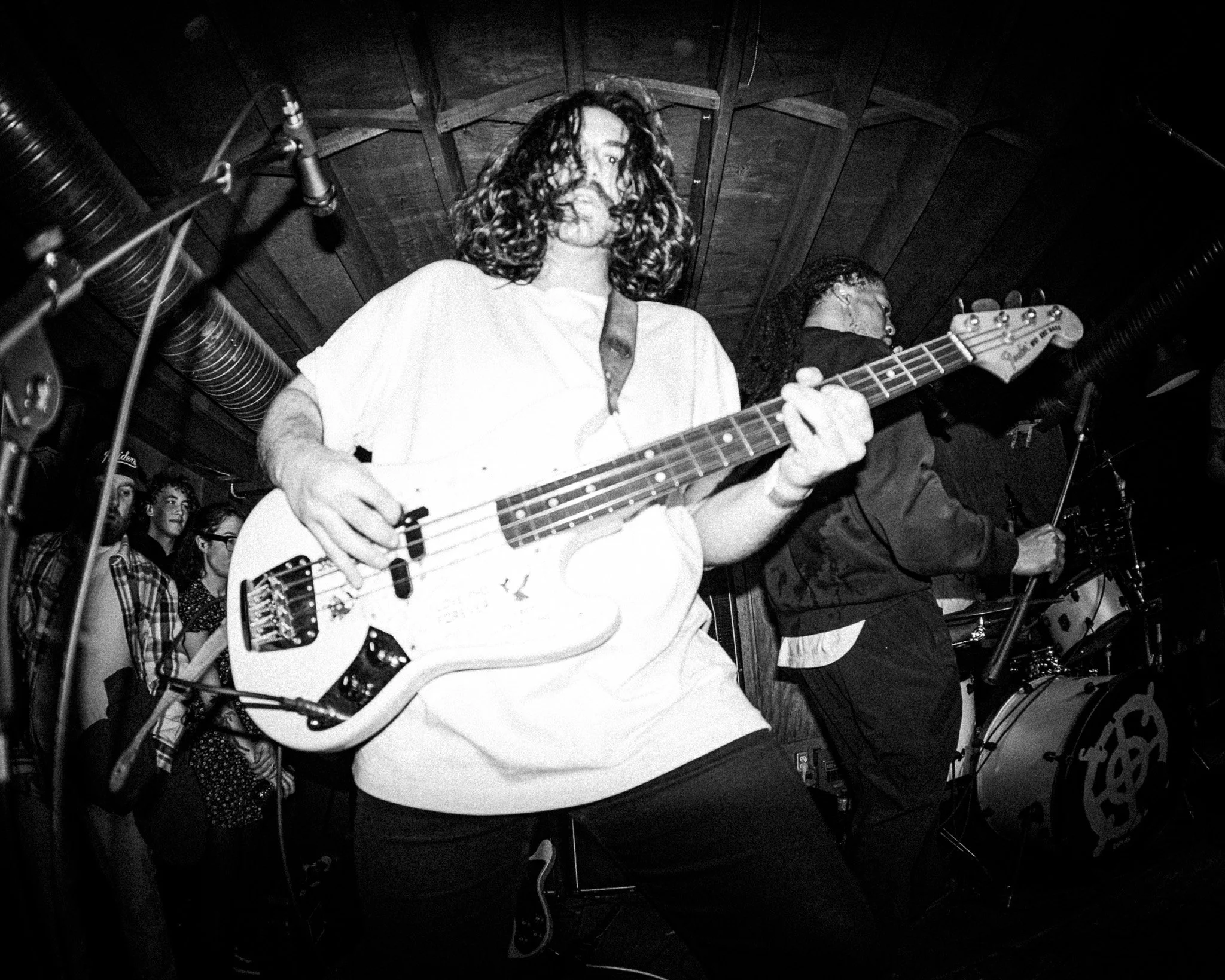 A black and white photo of a musician playing an electric bass guitar on stage, surrounded by a small crowd of fans.