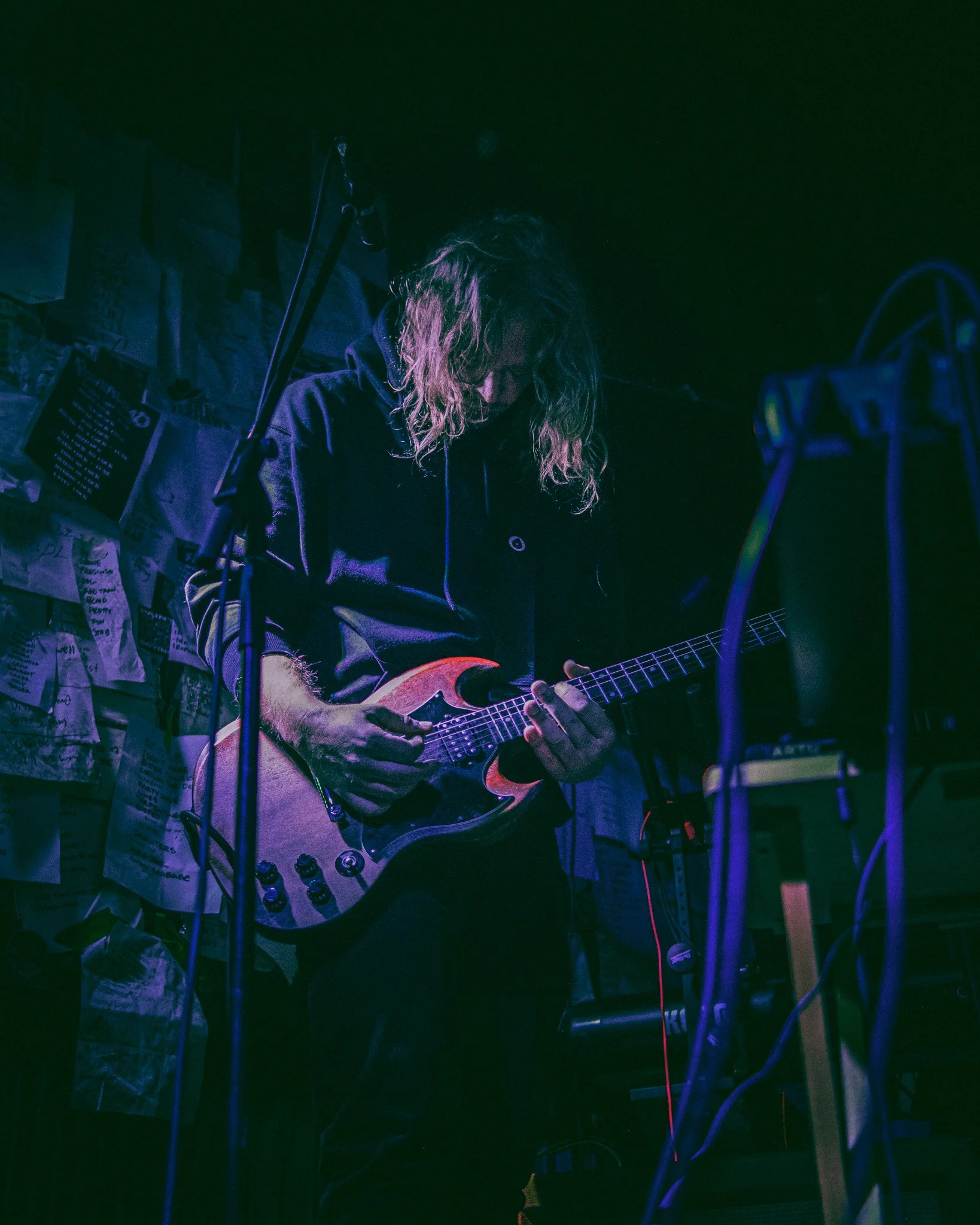 A person playing an electric guitar on stage, with a wall covered in notes and pictures behind them.