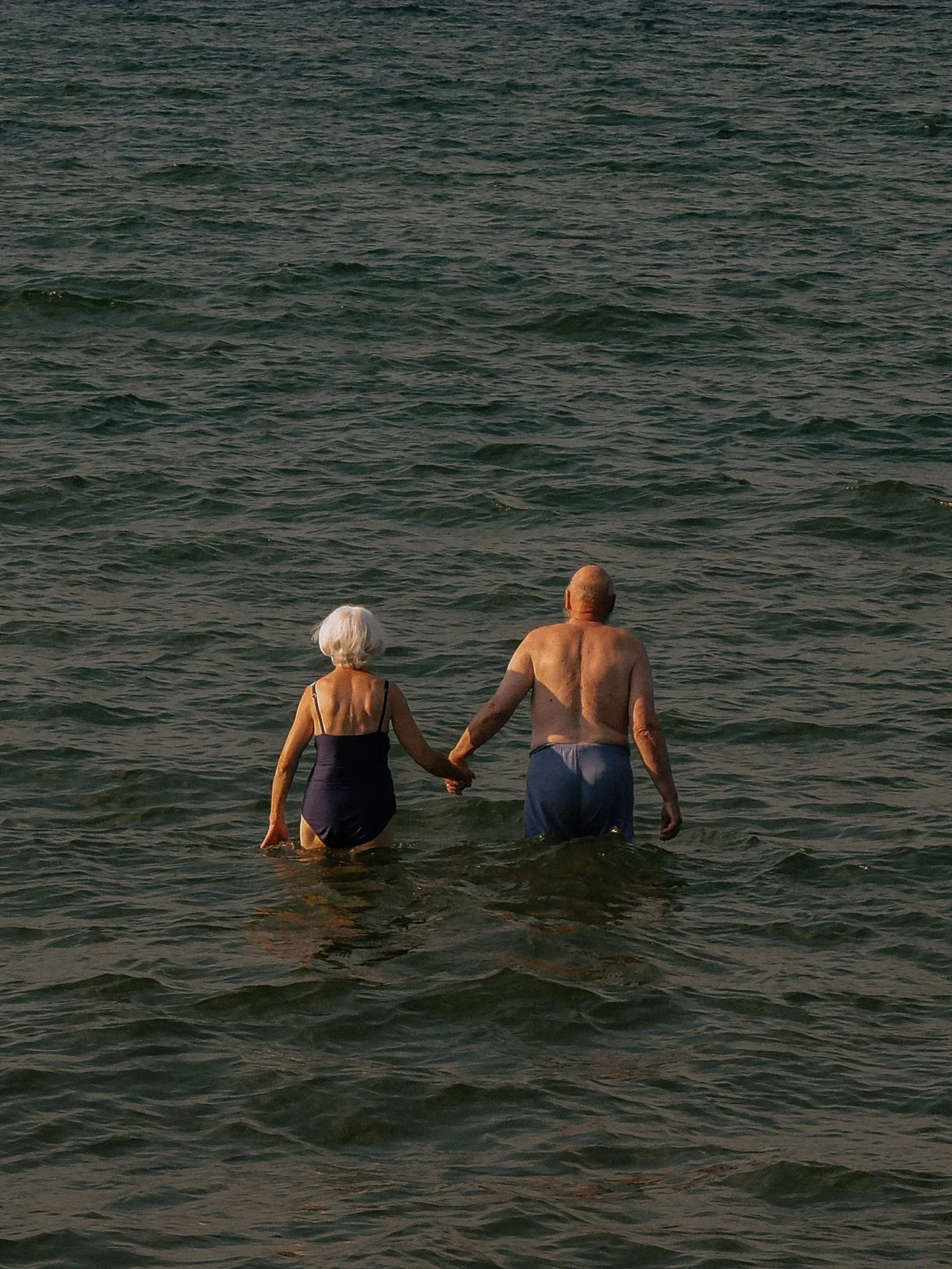 An elderly couple holds hands while wading in the ocean.