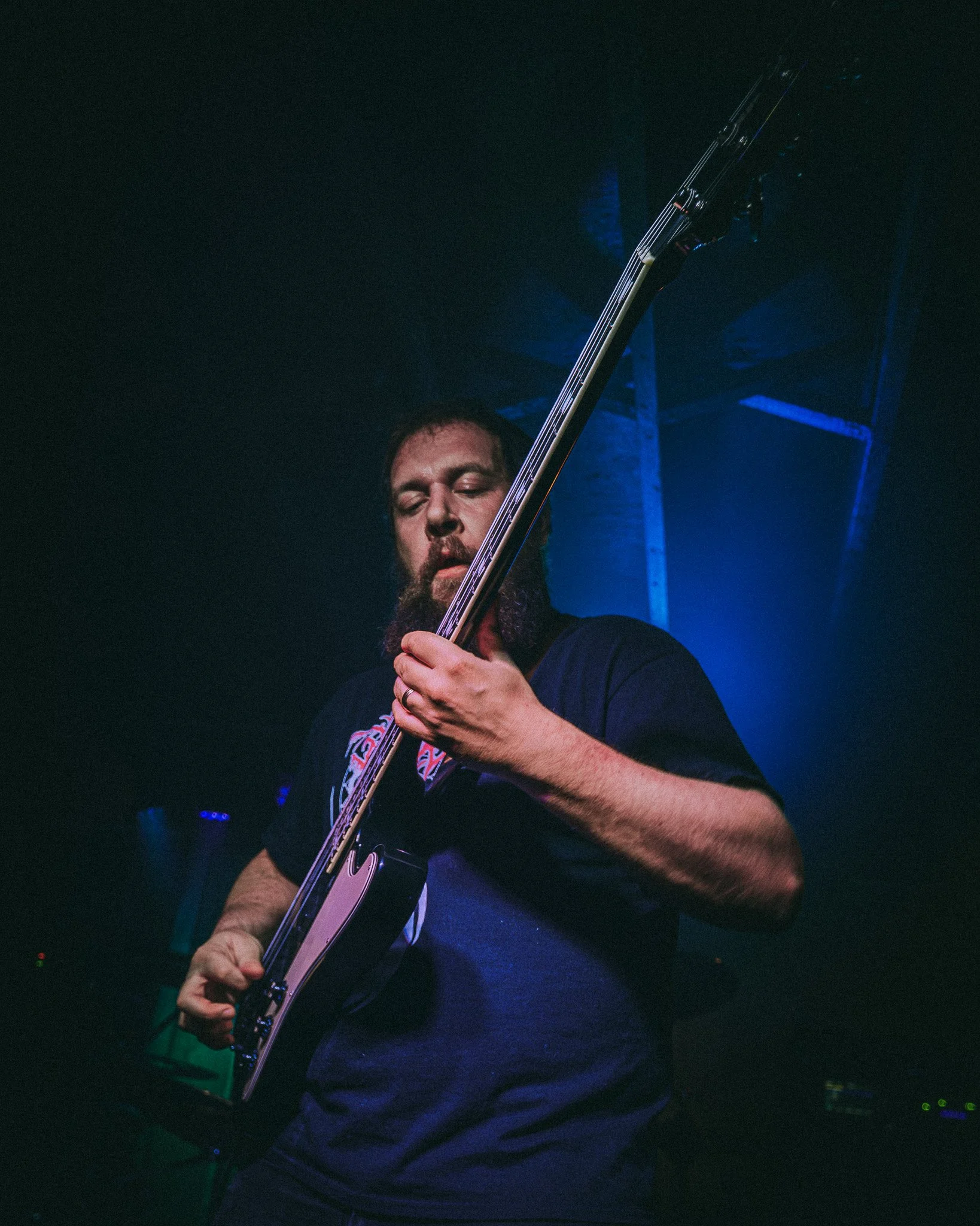 A man with a beard plays an electric guitar on stage in a dimly lit setting with blue and purple lighting.