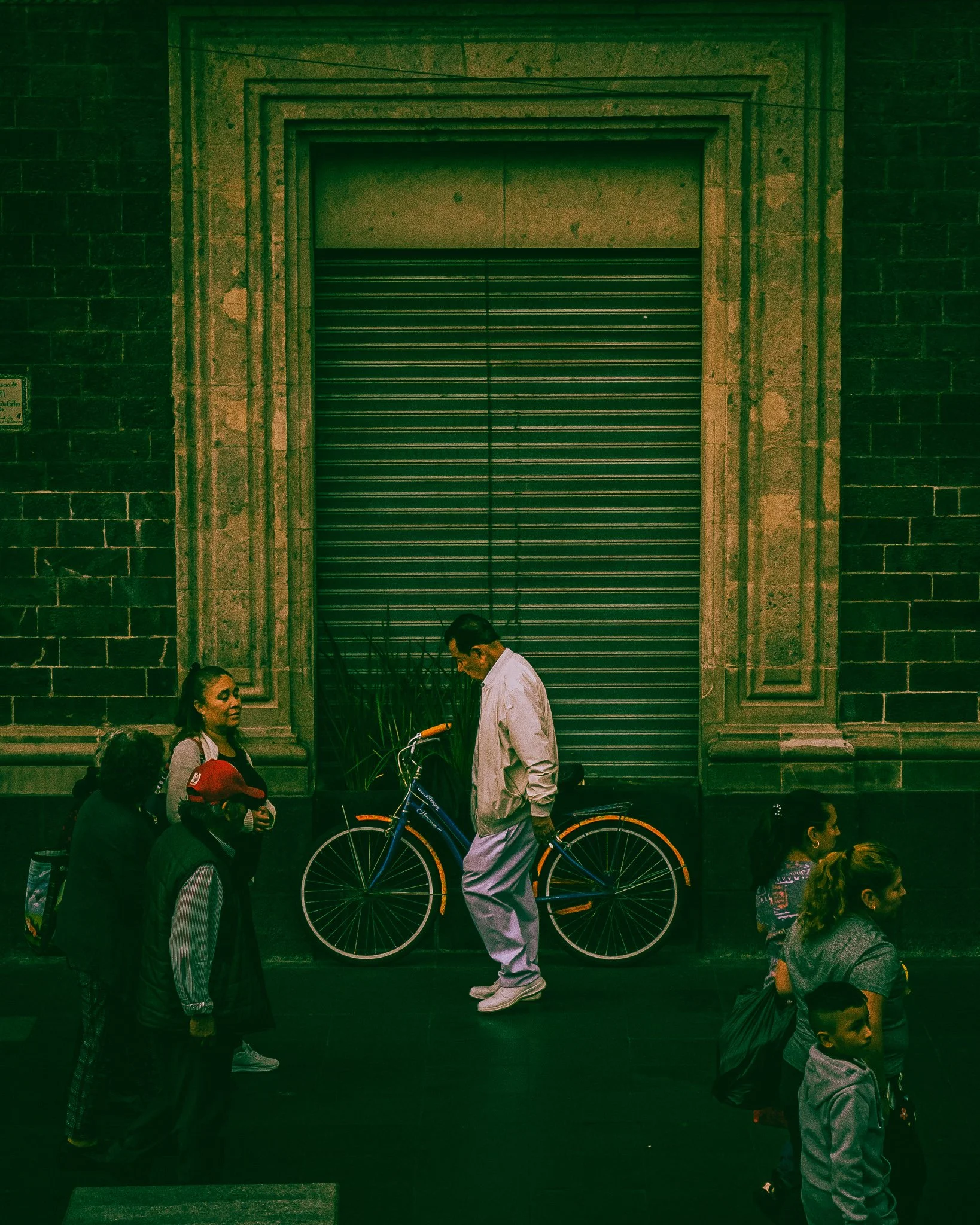 Man with bicycle on city street with several pedestrians, stone building with closed metal shutter in the background.