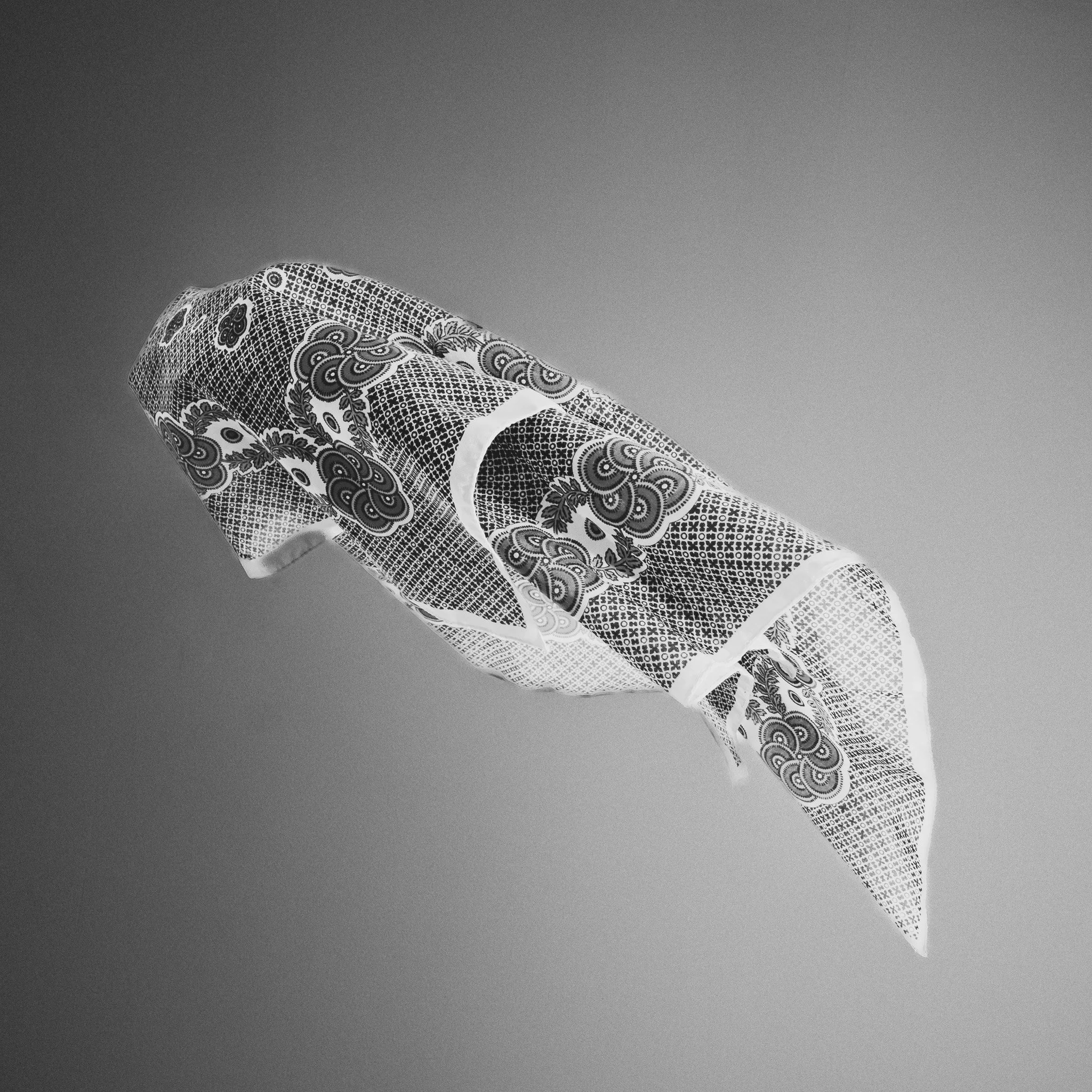 A bandana with intricate black and white paisley and geometric patterns, floating against a neutral gray background.