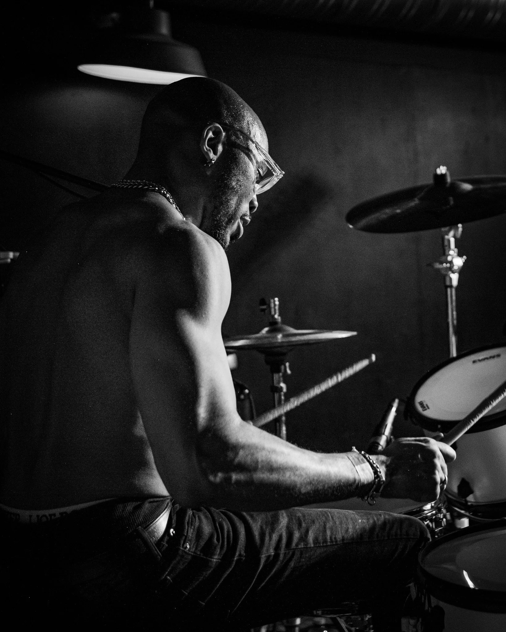 A shirtless man with glasses and jewelry playing the drums in a dimly lit setting.