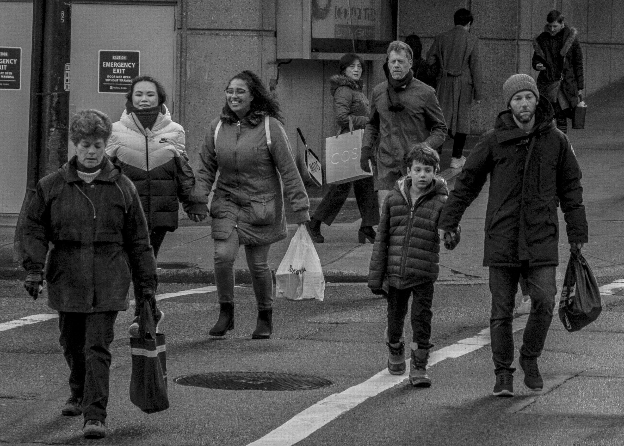 Group of people walking across a city street in cold weather. Adults and children wearing winter coats, some carrying shopping bags.