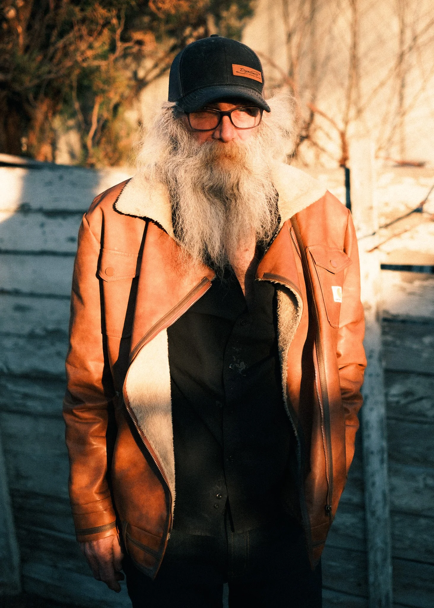 A man with long gray hair and a beard wearing glasses, a black cap, black shirt, and a tan Carhartt jacket standing outdoors at sunset.