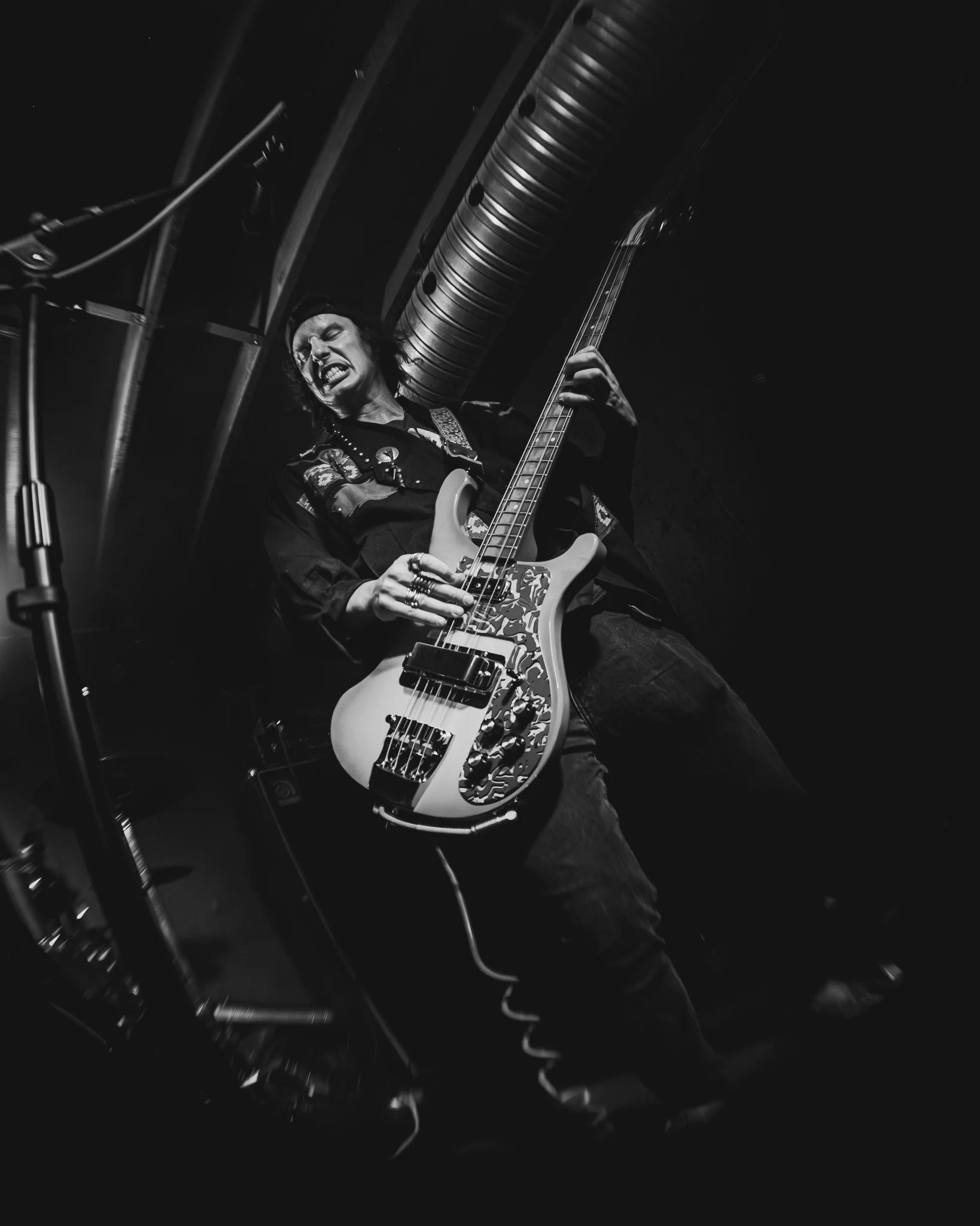Black and white photo of a musician playing an electric guitar passionately, with an intense facial expression, in a dimly lit setting.
