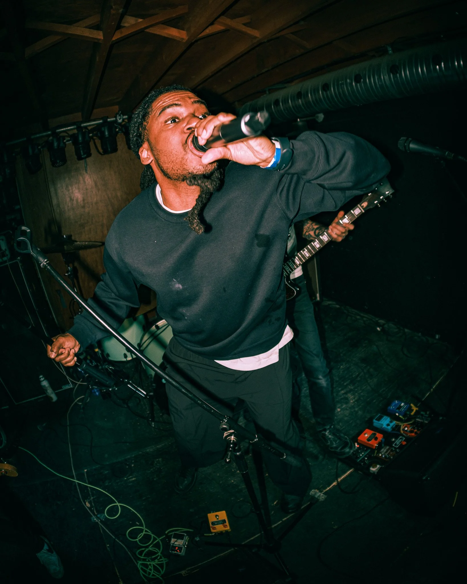 A male vocalist singing into a microphone on stage with a guitarist in the background, stage equipment, and pedal effects on the floor.