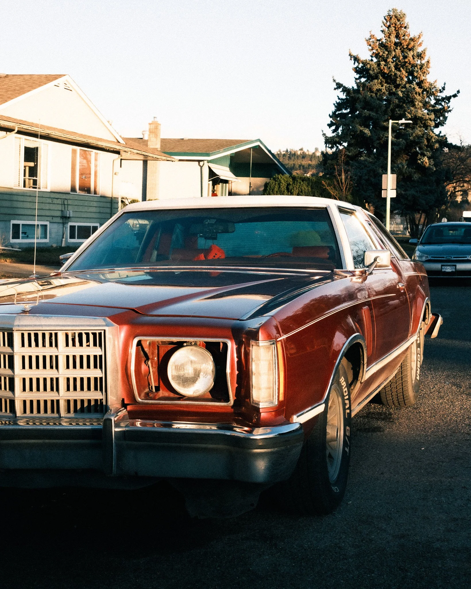 A vintage red car with a missing headlight parked on a residential street during sunset.