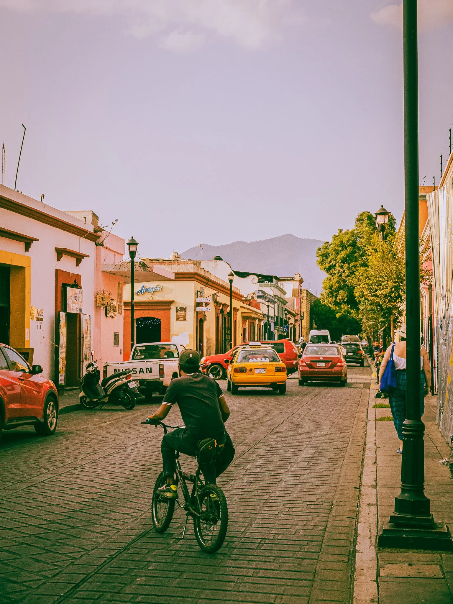 A person riding a bicycle on a cobblestone street lined with colorful buildings, cars, and street lamps, with mountains in the background.