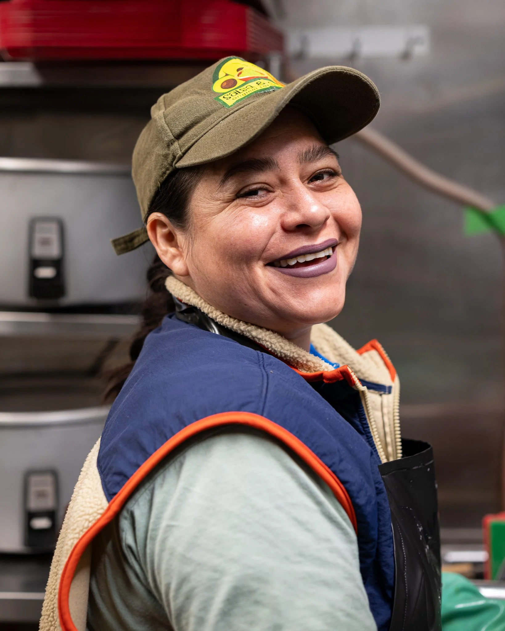 A smiling woman wearing a green cap, a blue vest, and a beige fleece-lined shirt, standing in a kitchen environment.