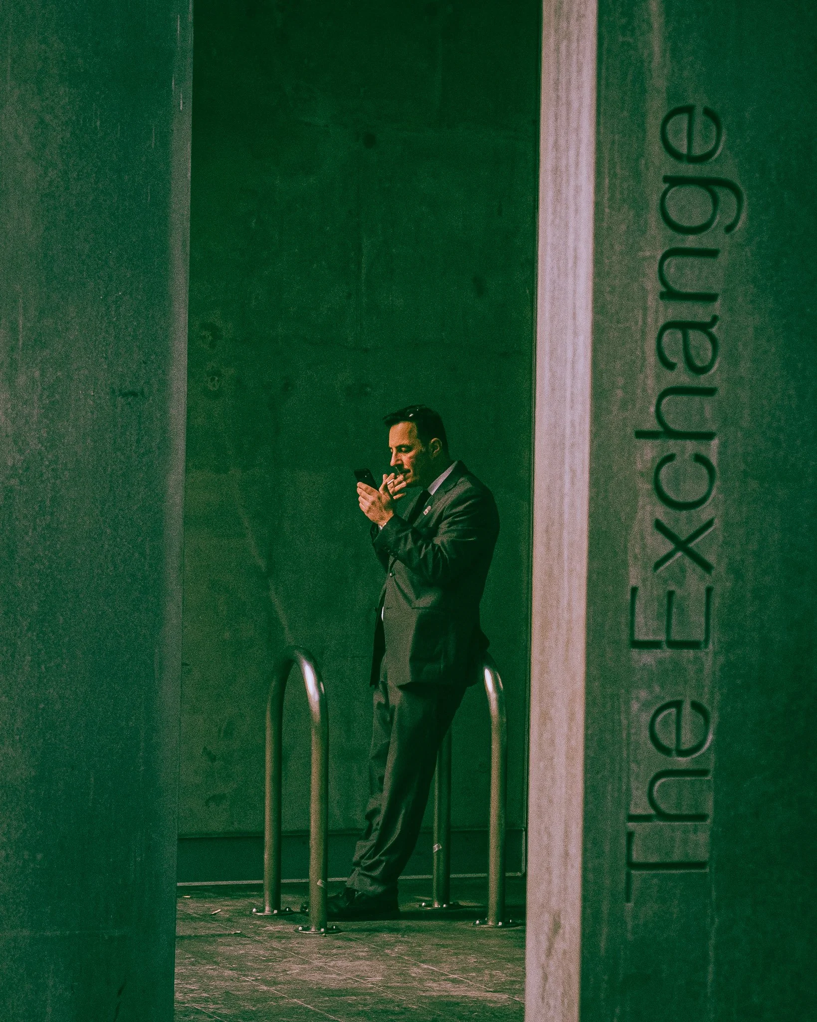 A man in a suit standing outside the entrance of a building, looking at his phone. The building has a sign reading 'The Exchange'.