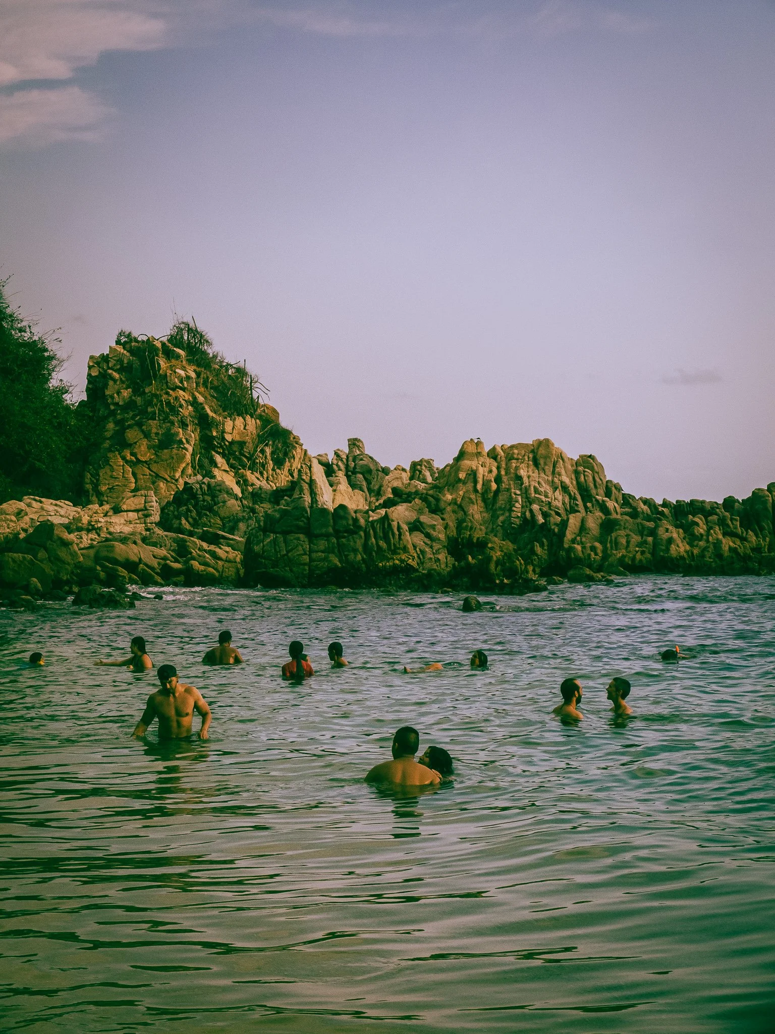 People swimming and wading in the ocean near rocky cliffs under a clear sky.