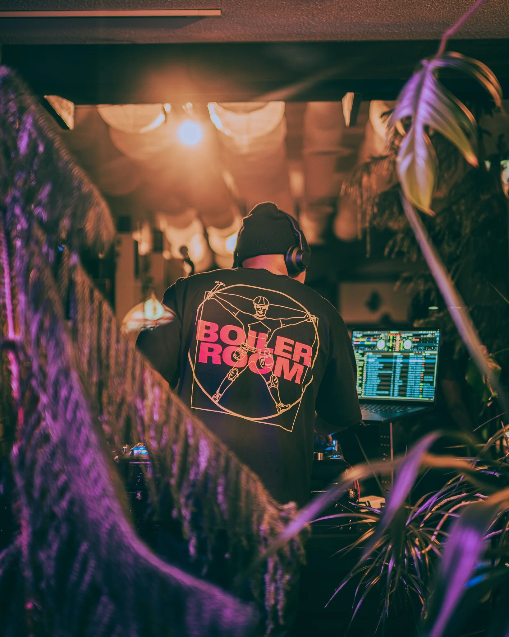 A DJ wearing headphones and a black hoodie with a skeleton graphic and 'BOILER ROOM' logo, performing at a nightclub with colorful lights and a mixing console.