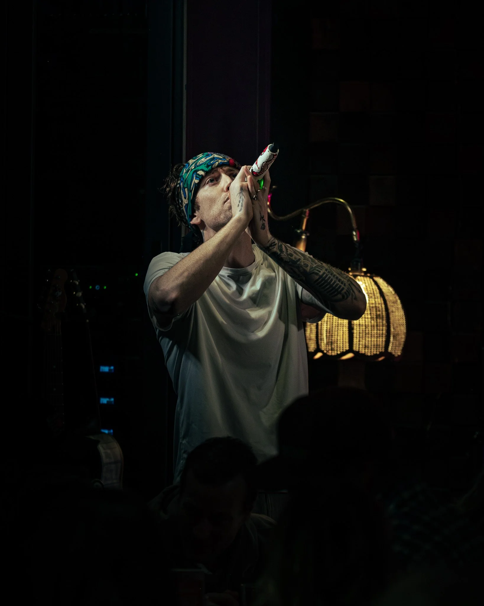 A man singing into a microphone on stage, wearing a patterned headband and tattoos on his arms.